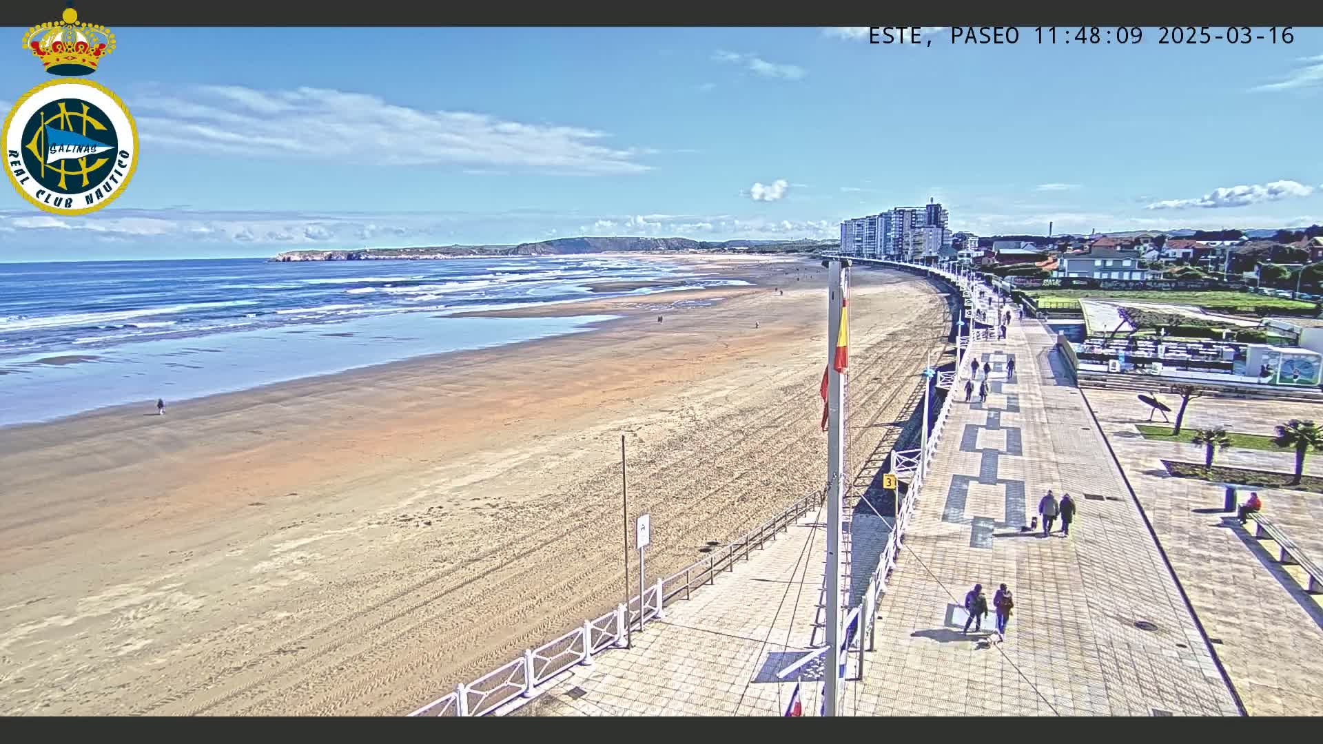 A wide sandy beach with small waves under a mostly sunny sky is bordered by a paved boardwalk with several people walking along it and buildings in the background.