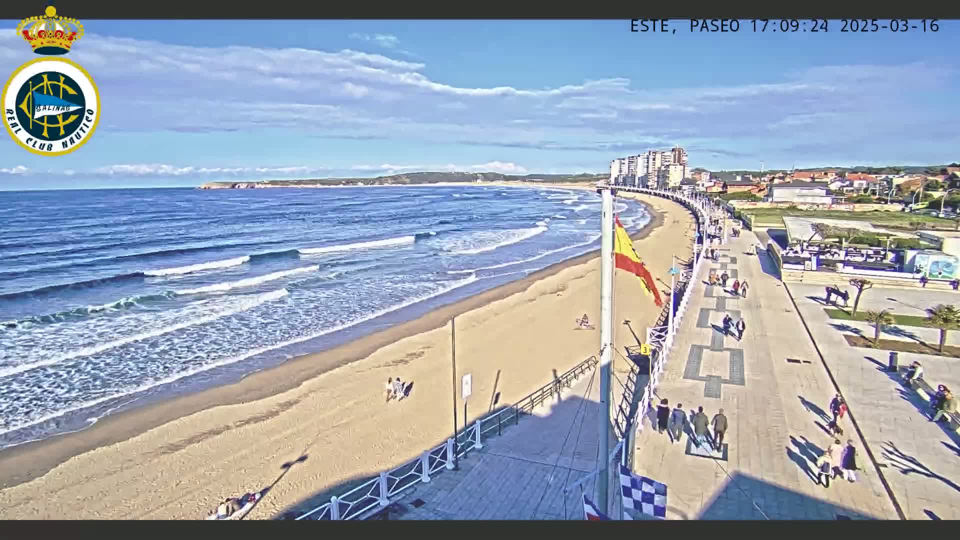 A sandy beach with gentle waves under a mostly sunny sky is bordered by a paved promenade with people walking along it, and buildings are visible in the background.