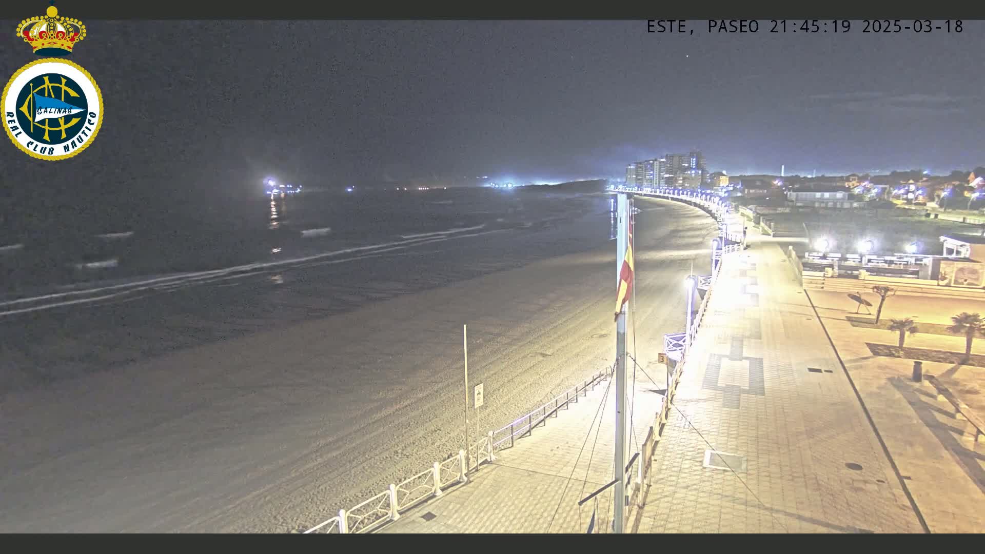 A nighttime view of a sandy beach and boardwalk, with calm ocean waves and buildings visible in the distance under a dark sky.