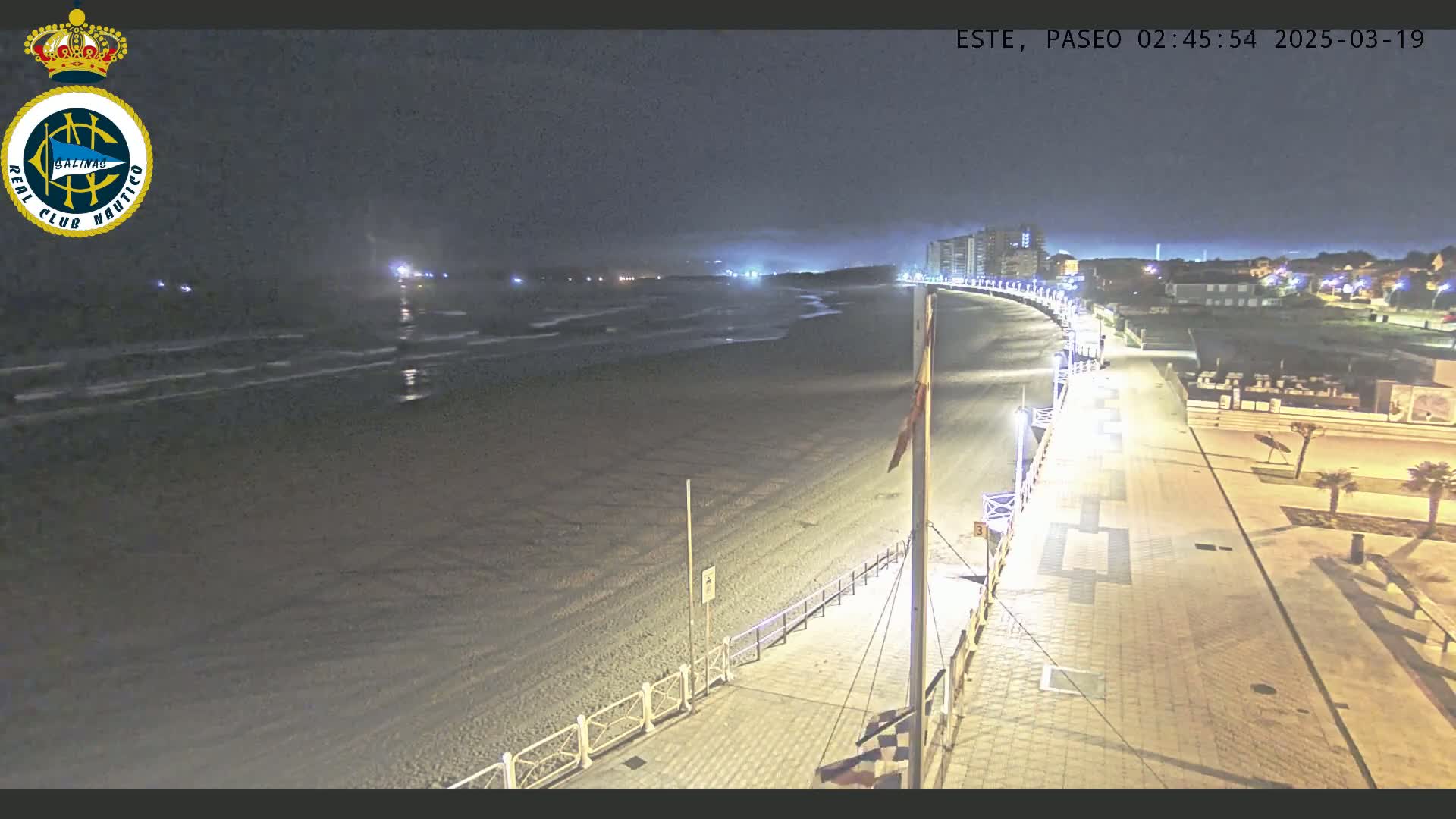 A nighttime view of a mostly empty beach with gentle waves, a paved walkway, and buildings in the distance under a dark sky.