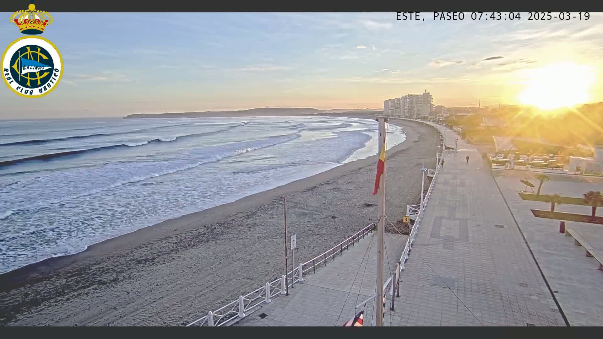 A sandy beach with gently rolling waves is seen at sunrise next to a paved walkway and buildings.