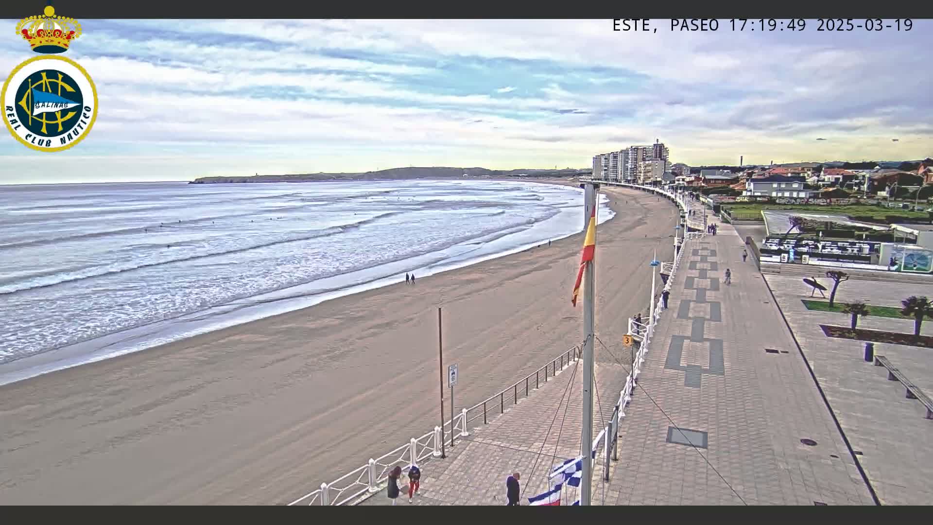 A sandy beach with gentle waves under a partly cloudy sky is bordered by a paved walkway leading to buildings and a promenade.