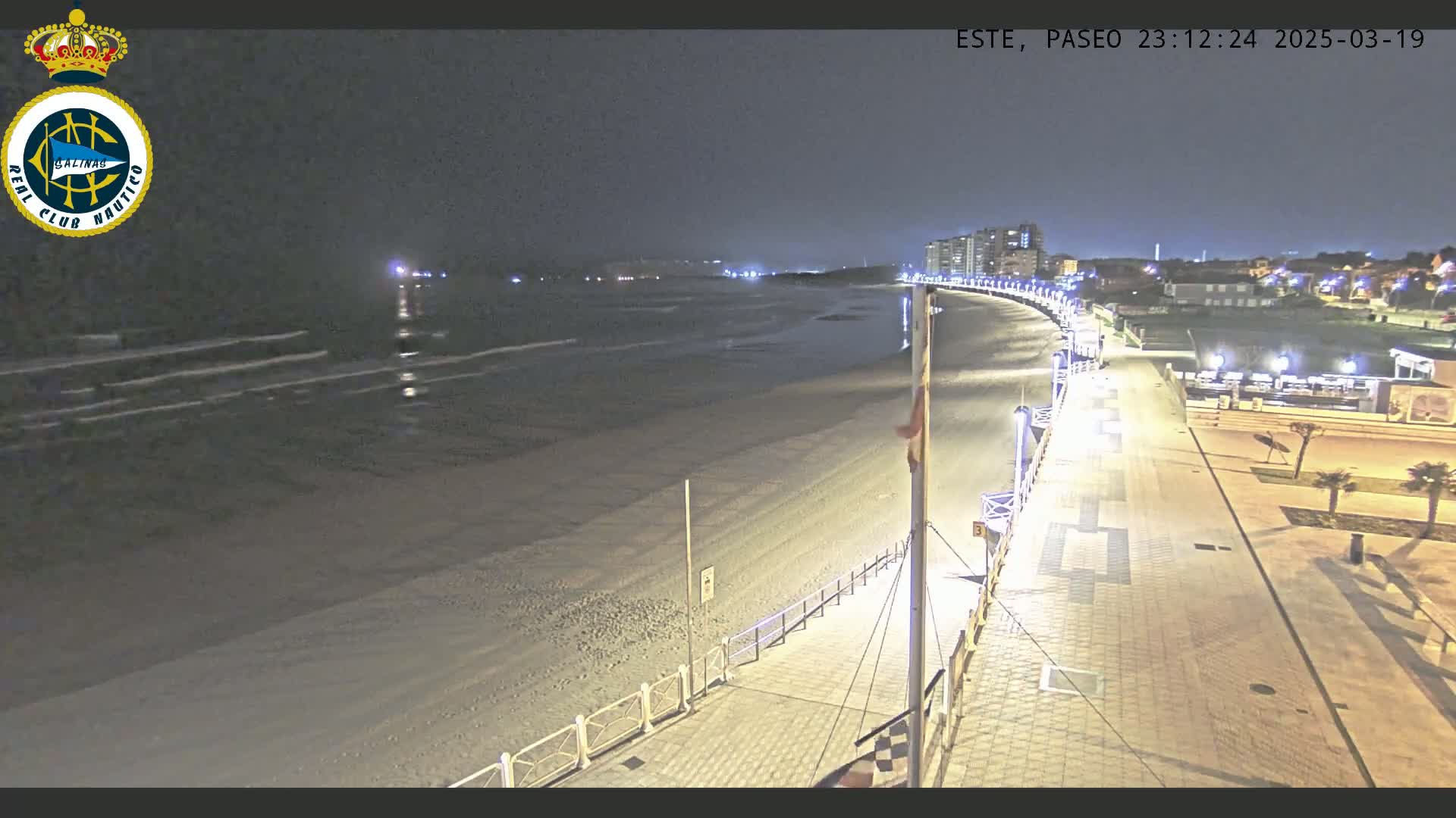 A nighttime view of a mostly empty sandy beach, gently lapping waves, and a well-lit boardwalk curving along the shore under a dark sky.