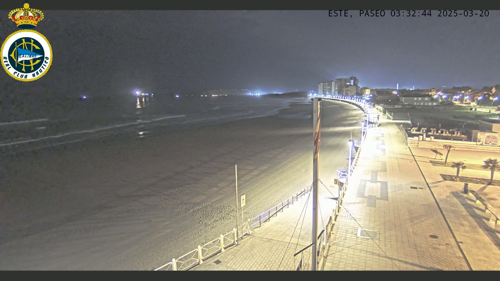 A nighttime view of a mostly empty beach with a paved walkway and lights, gentle waves lapping the shore, and buildings visible in the distance.