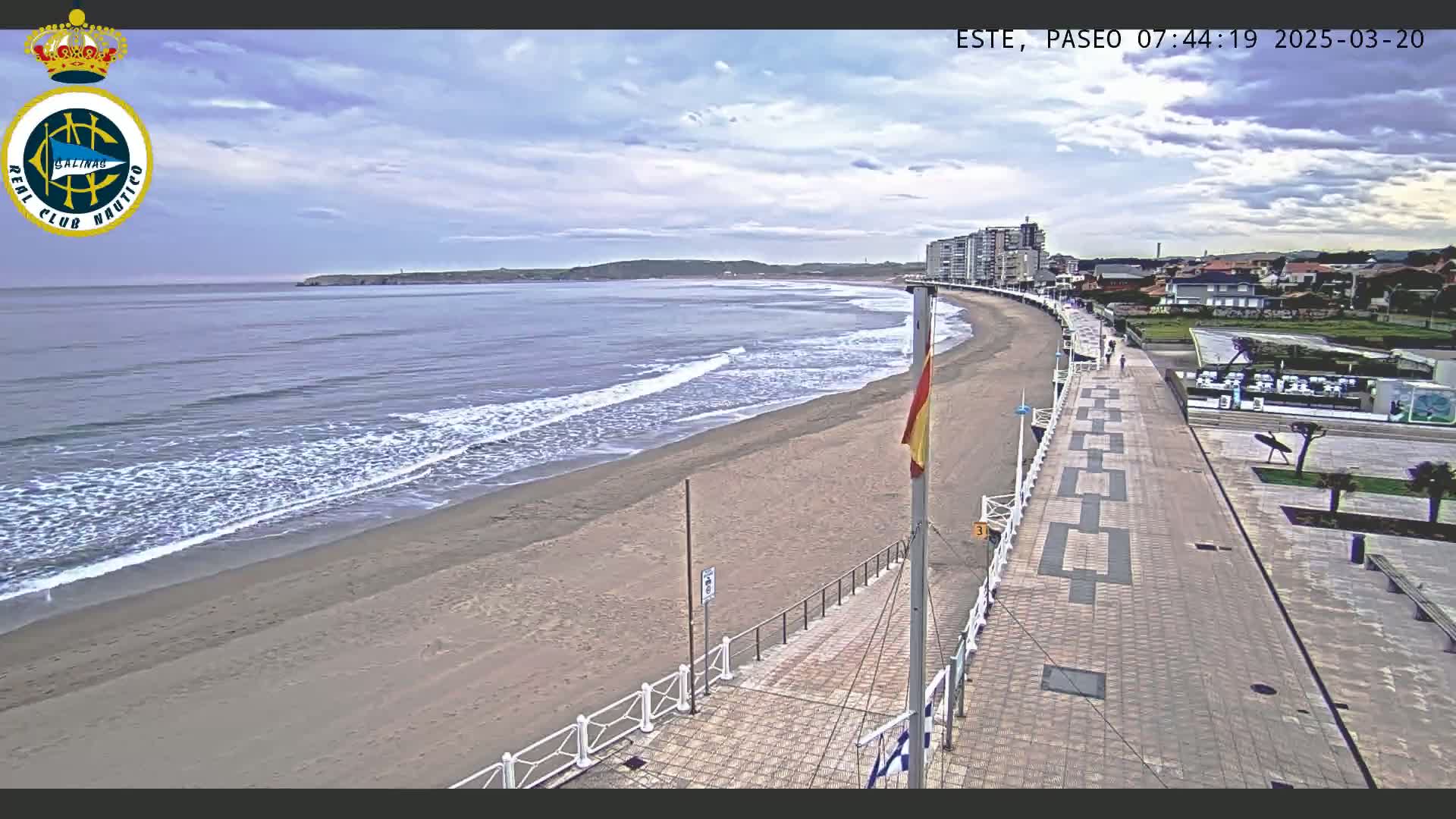 A sandy beach with gentle waves laps the shore under a mostly cloudy sky, next to a paved promenade with buildings in the background.