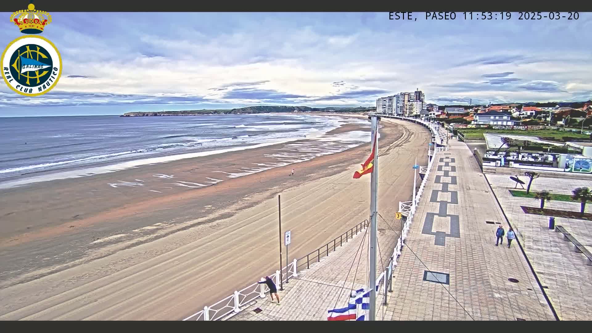 A sandy beach with gentle waves under a partly cloudy sky is bordered by a paved walkway and buildings.