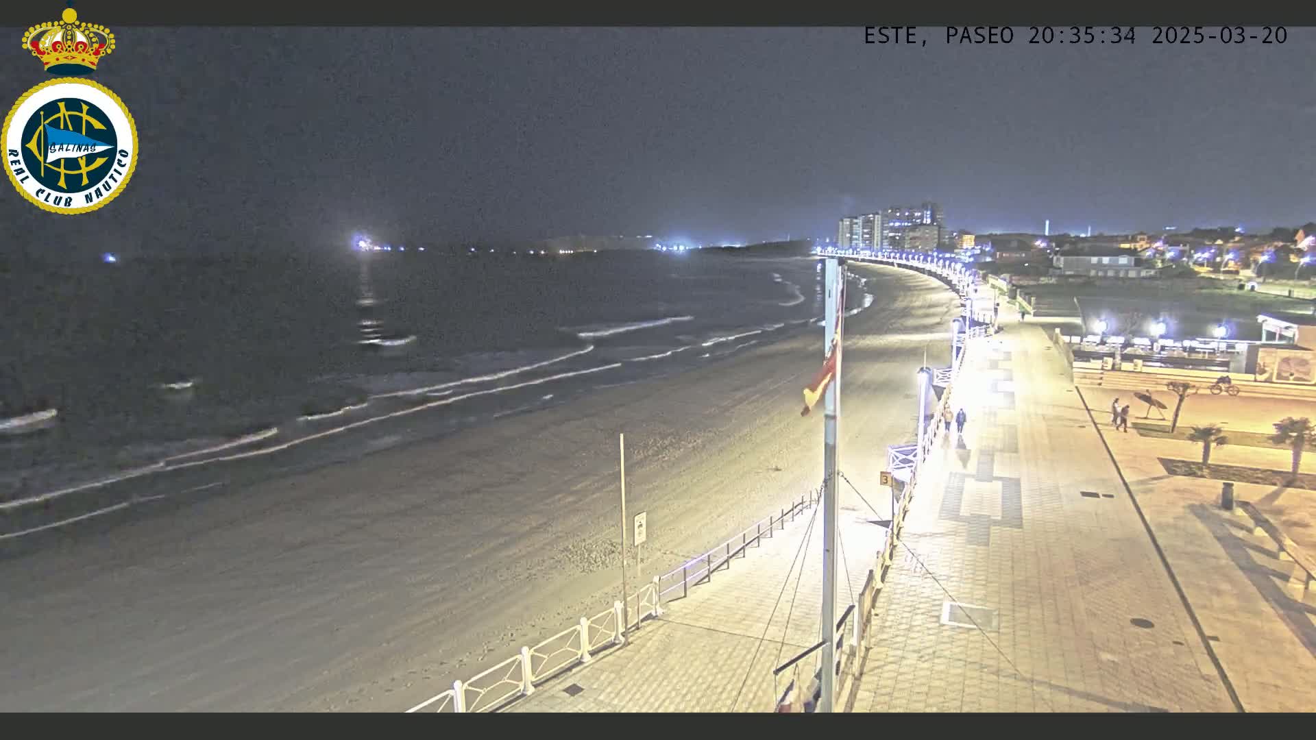 A nighttime view of a beach with gentle waves, a paved boardwalk lined with lights, and a few people strolling along the shore under a dark sky.