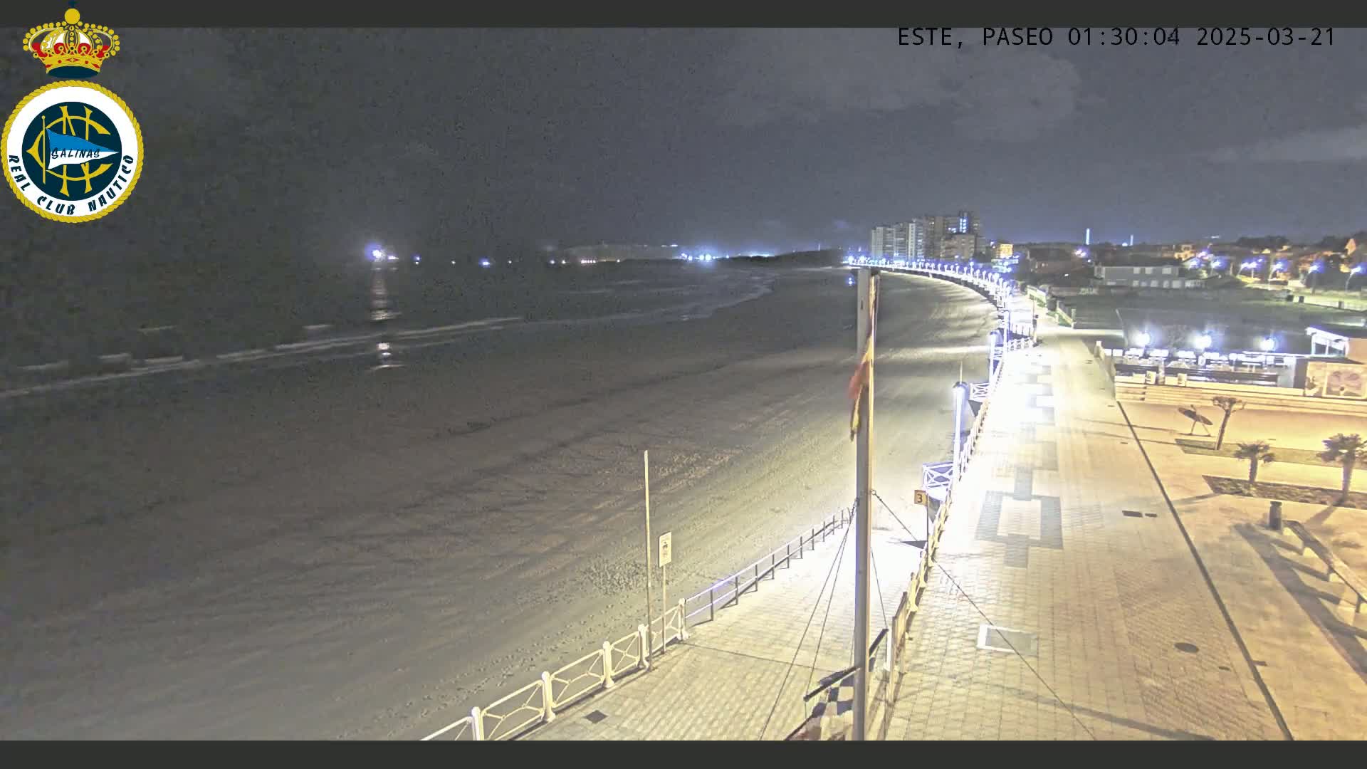 A nighttime, high-angle view of a mostly empty sandy beach and a well-lit boardwalk curving along the shore, with buildings visible in the distance.
