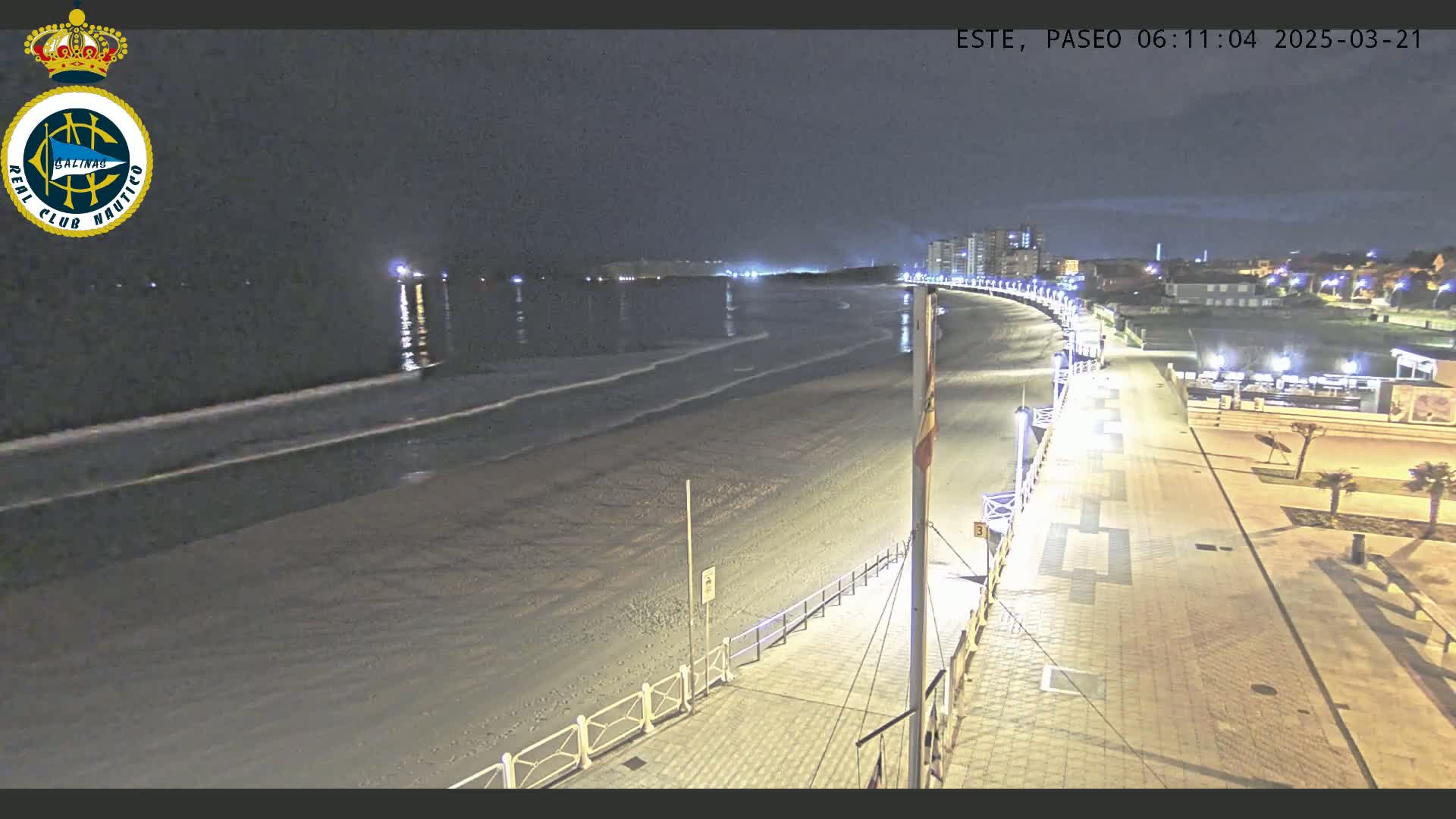 A nighttime view of a mostly empty beach and adjacent boardwalk, with gentle waves lapping the shore and distant lights illuminating buildings.