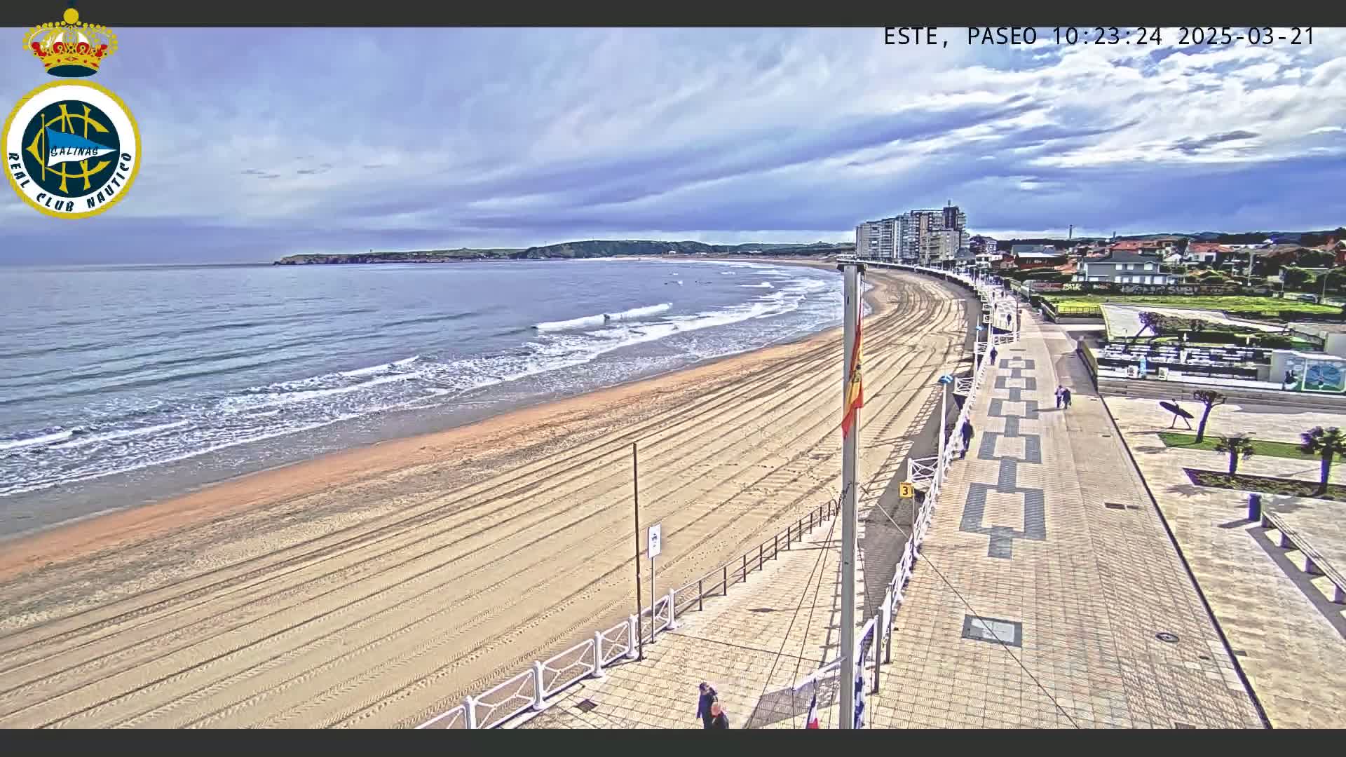 A sandy beach with gentle waves stretches alongside a paved boardwalk, under a partly cloudy sky.