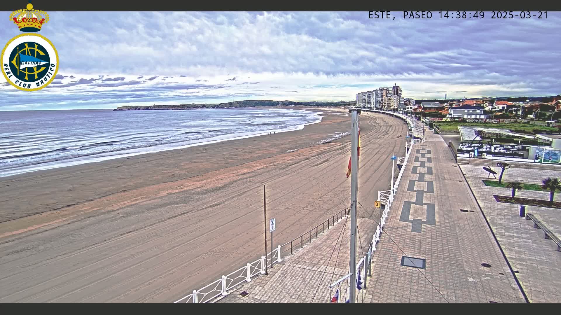 An overcast day shows a sandy beach with small waves, a paved boardwalk, and buildings in the background.
