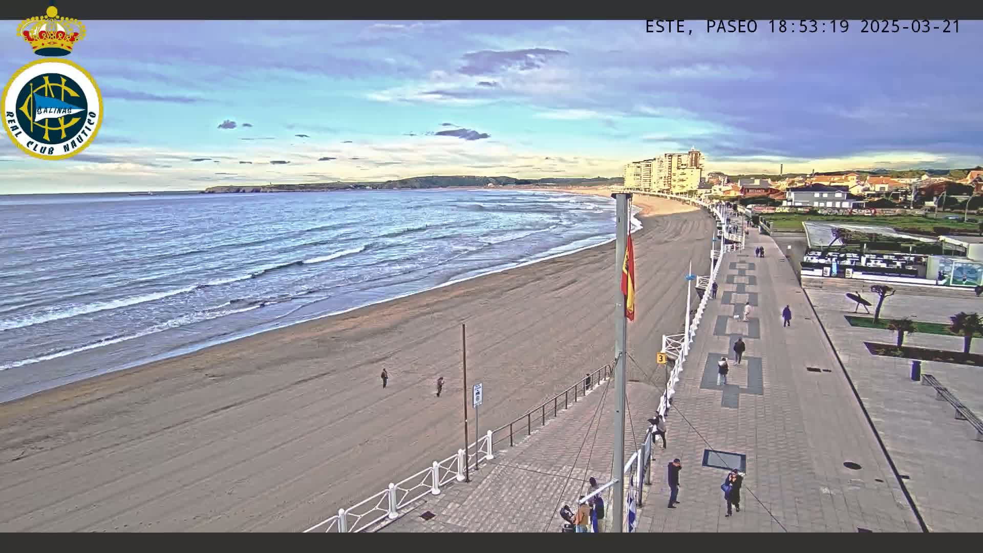 A sandy beach with gentle waves under a partly cloudy sky is viewed from above, showing a paved walkway with people strolling along it next to buildings.