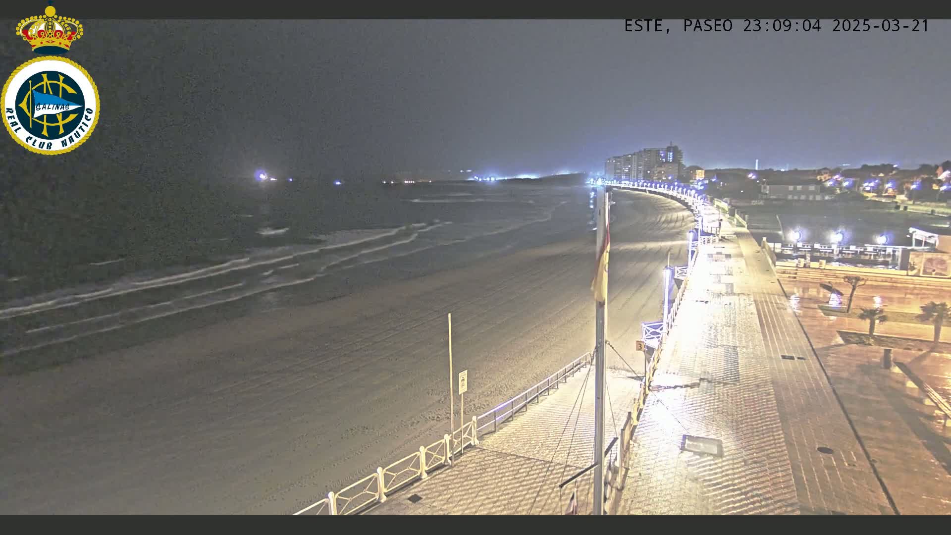 A nighttime view of a sandy beach with gentle waves, a paved boardwalk, and buildings in the distance under a dark, possibly cloudy sky.