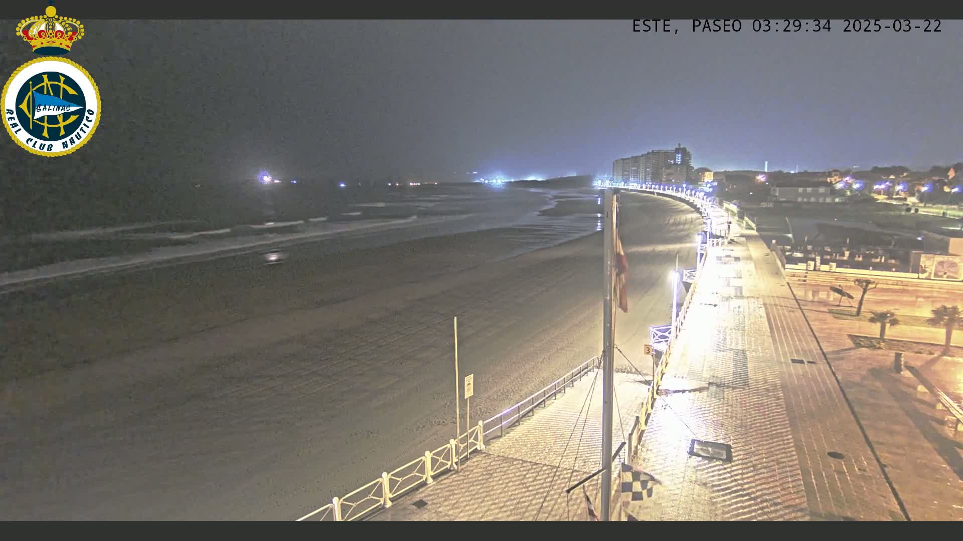 A nighttime view of a mostly empty beach and boardwalk, with gentle waves lapping the shore under a dark sky.