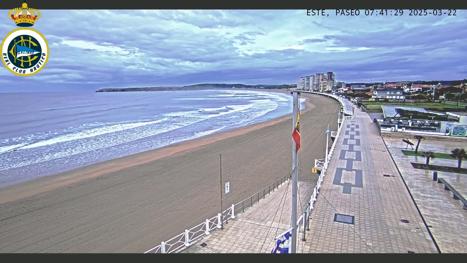 A sandy beach with gentle waves under a mostly cloudy sky is bordered by a paved walkway and buildings.