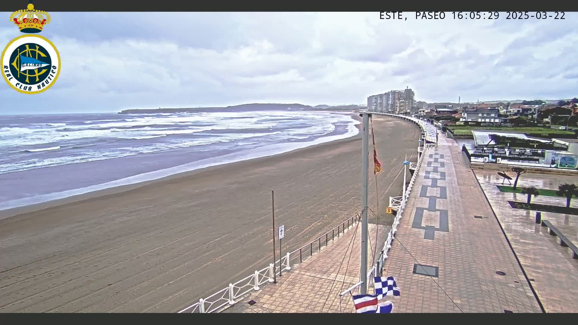 A sandy beach with moderate waves under an overcast sky is adjacent to a paved walkway and buildings.