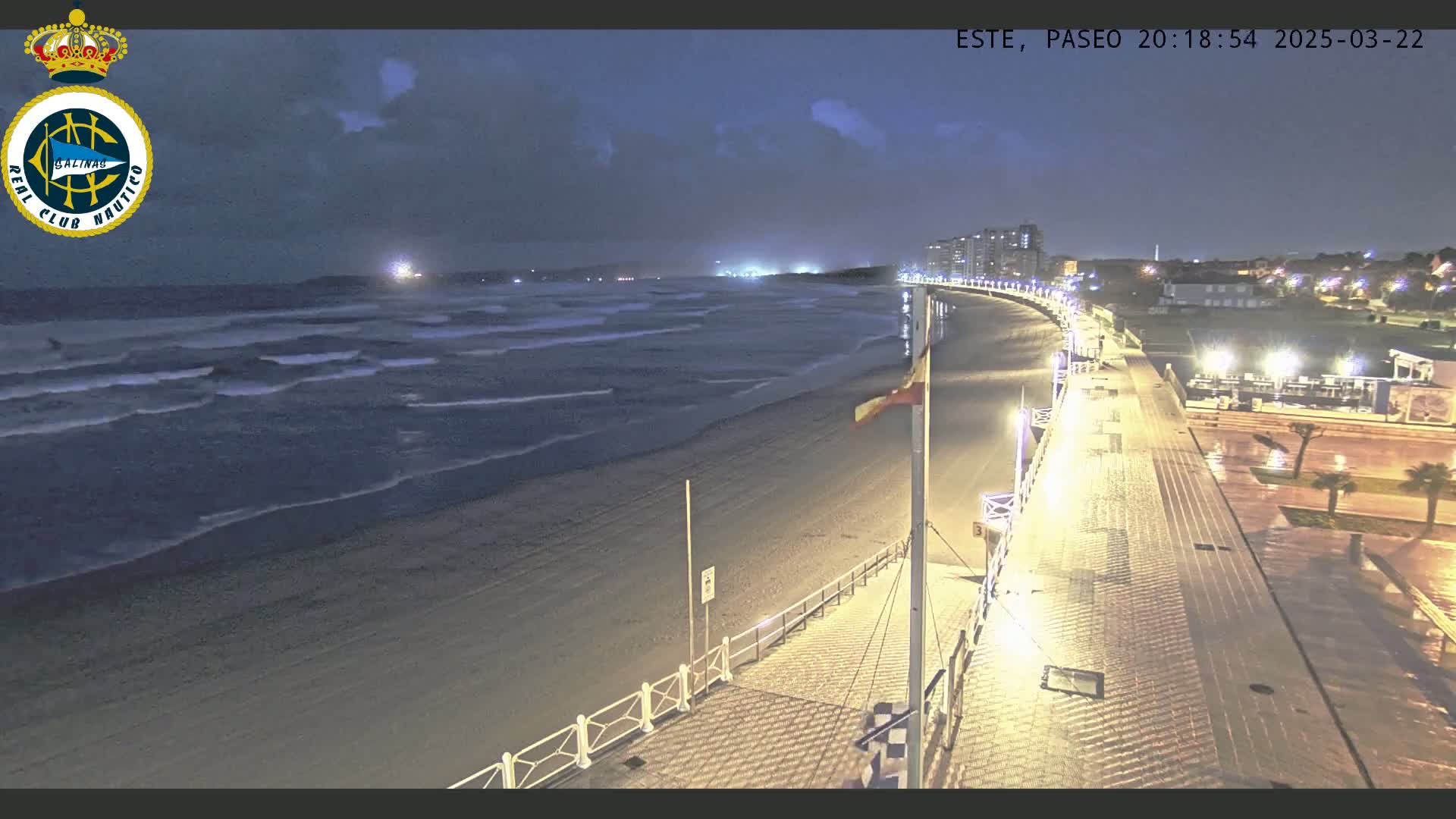 A nighttime view of a beach with moderate waves under a partly cloudy sky, showing a boardwalk with lights extending from the sandy shore to buildings in the distance.