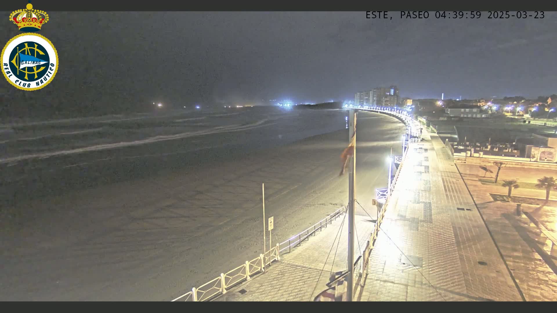 A nighttime view of a beach with gently rolling waves under a dark sky, showing a paved boardwalk and buildings in the distance.
