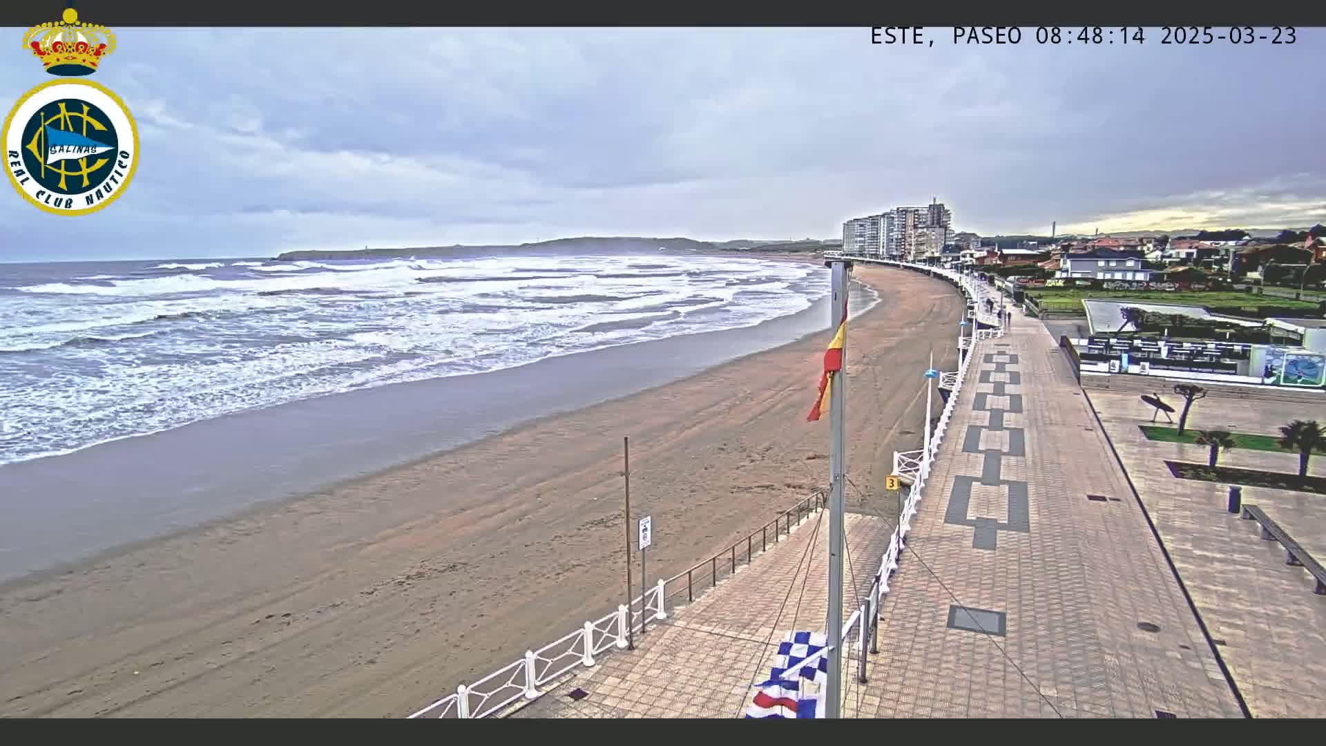 A sandy beach with moderate waves under an overcast sky is seen from above, alongside a paved boardwalk and buildings in the distance.