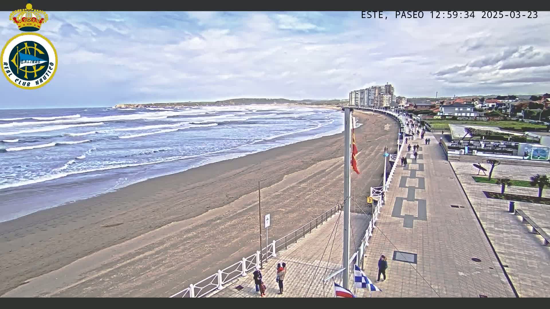 A wide sandy beach with moderate waves under a partly cloudy sky is bordered by a paved boardwalk with people walking, leading to buildings in the distance.