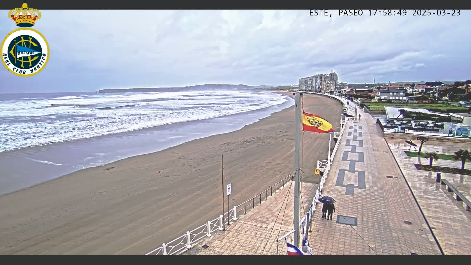 A sandy beach with moderate waves under an overcast sky is viewed from above a paved boardwalk, where two people with an umbrella are walking.