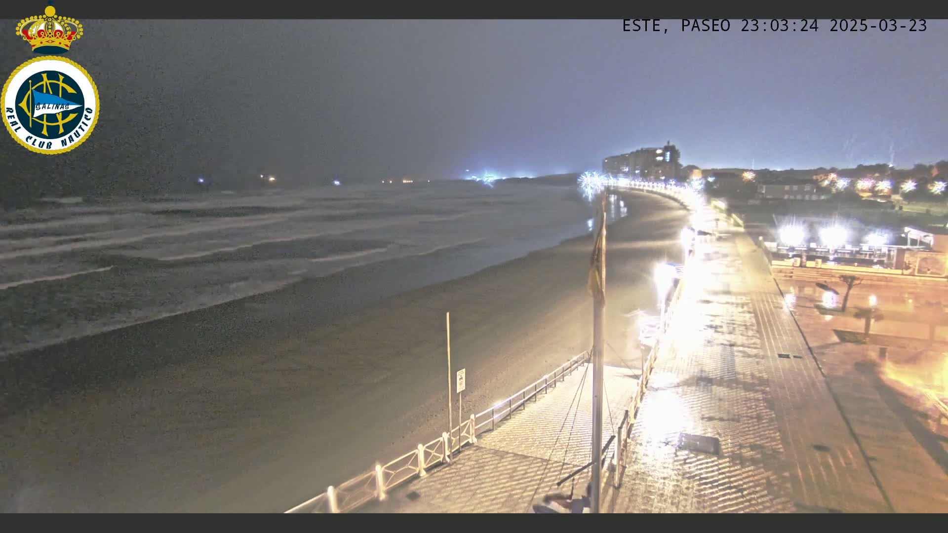 A nighttime view of a rain-soaked beach promenade with moderately sized waves crashing on the shore under a dark sky.