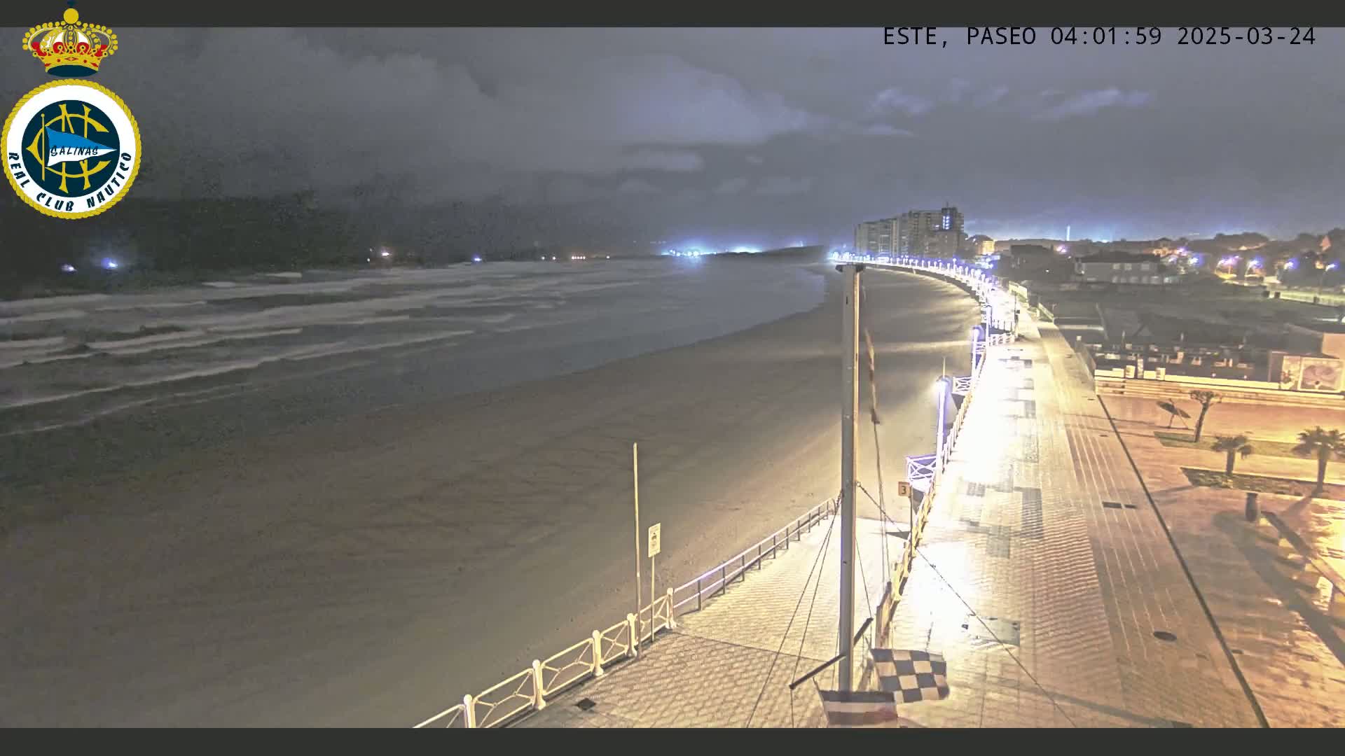 A nighttime view of a mostly empty beach with gently lapping waves, a boardwalk with lights, and buildings in the distance under a cloudy sky.