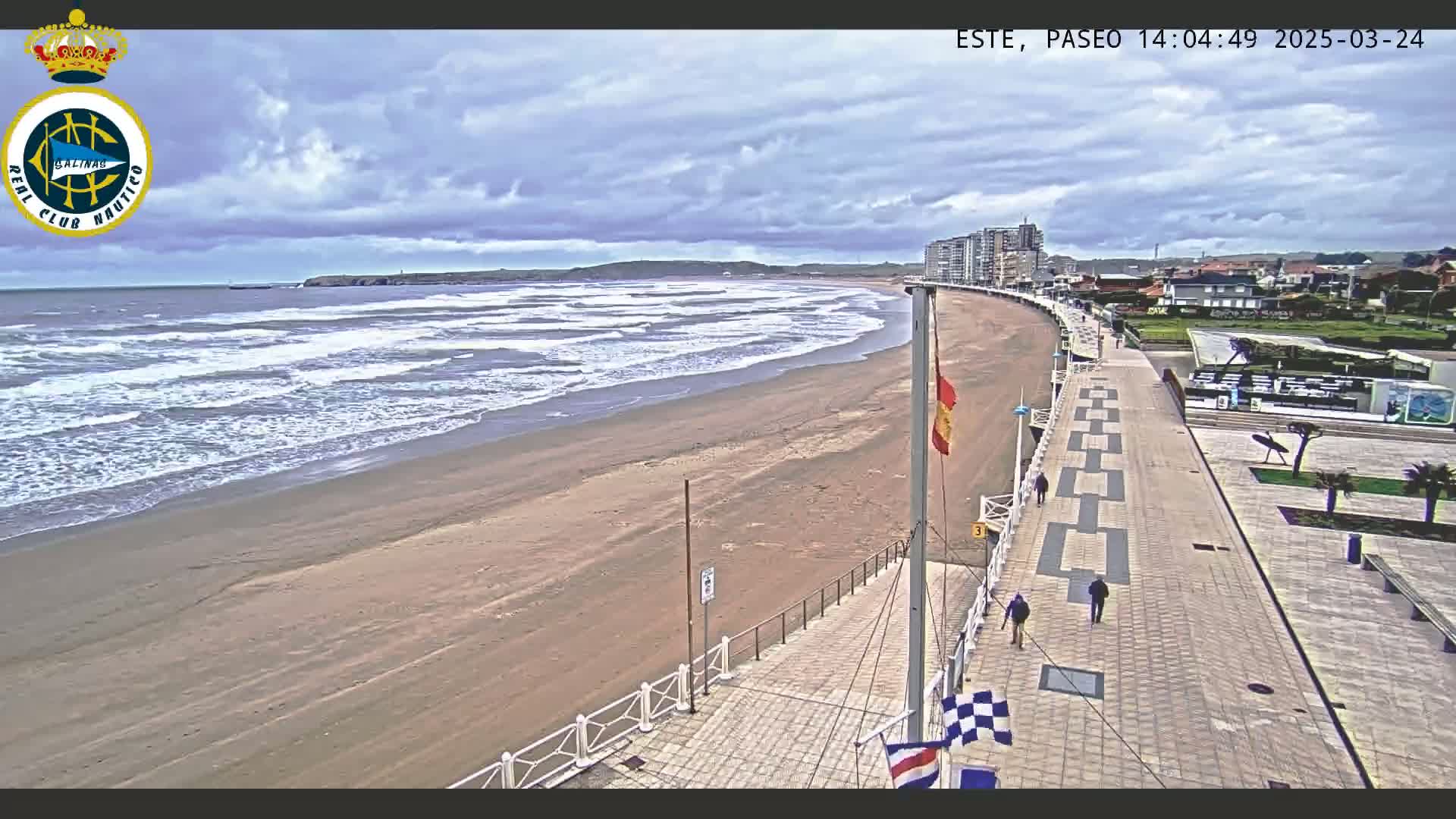 A sandy beach with moderate waves under a partly cloudy sky is visible from an elevated viewpoint overlooking a paved walkway and buildings.
