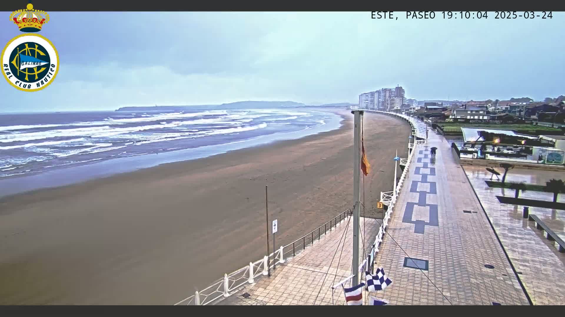 A wet sandy beach with gentle waves under a cloudy sky is visible from a high vantage point overlooking a paved walkway leading to buildings in the distance.