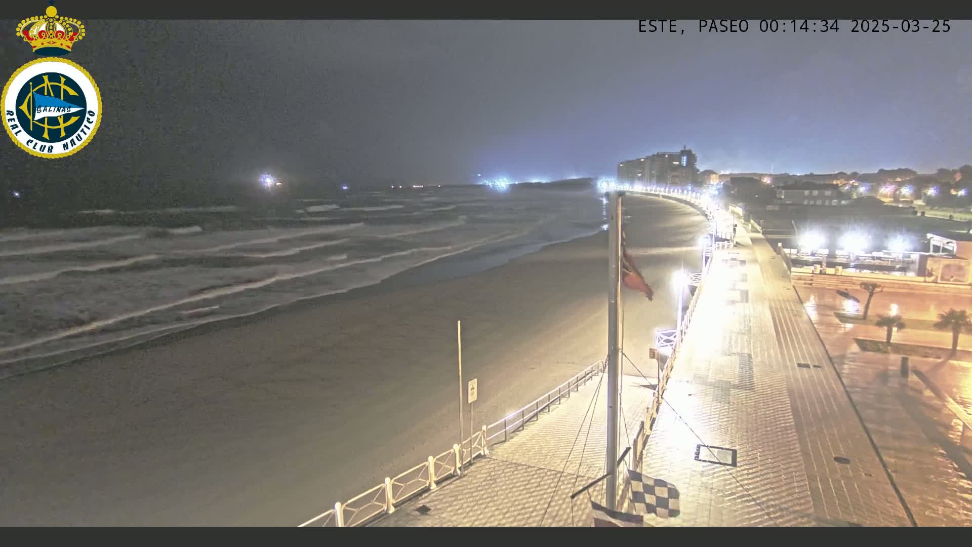 A nighttime view of a beach with gentle waves under a dark sky, showing a brightly lit boardwalk extending from the beach to buildings in the distance.