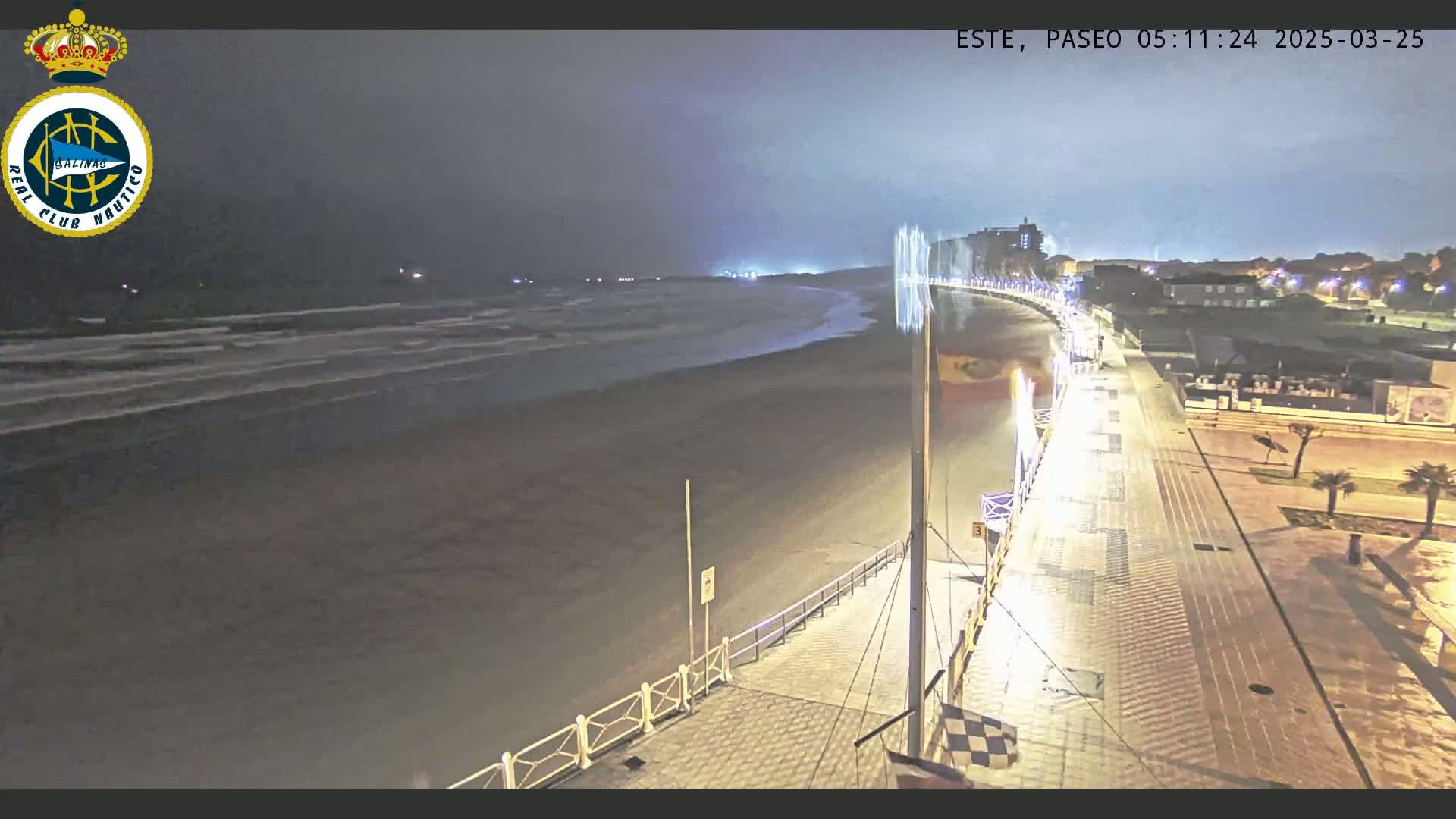 A nighttime view of a beach with dark, choppy waves under a cloudy sky, showing a boardwalk with lights stretching from the beach toward a town in the distance.