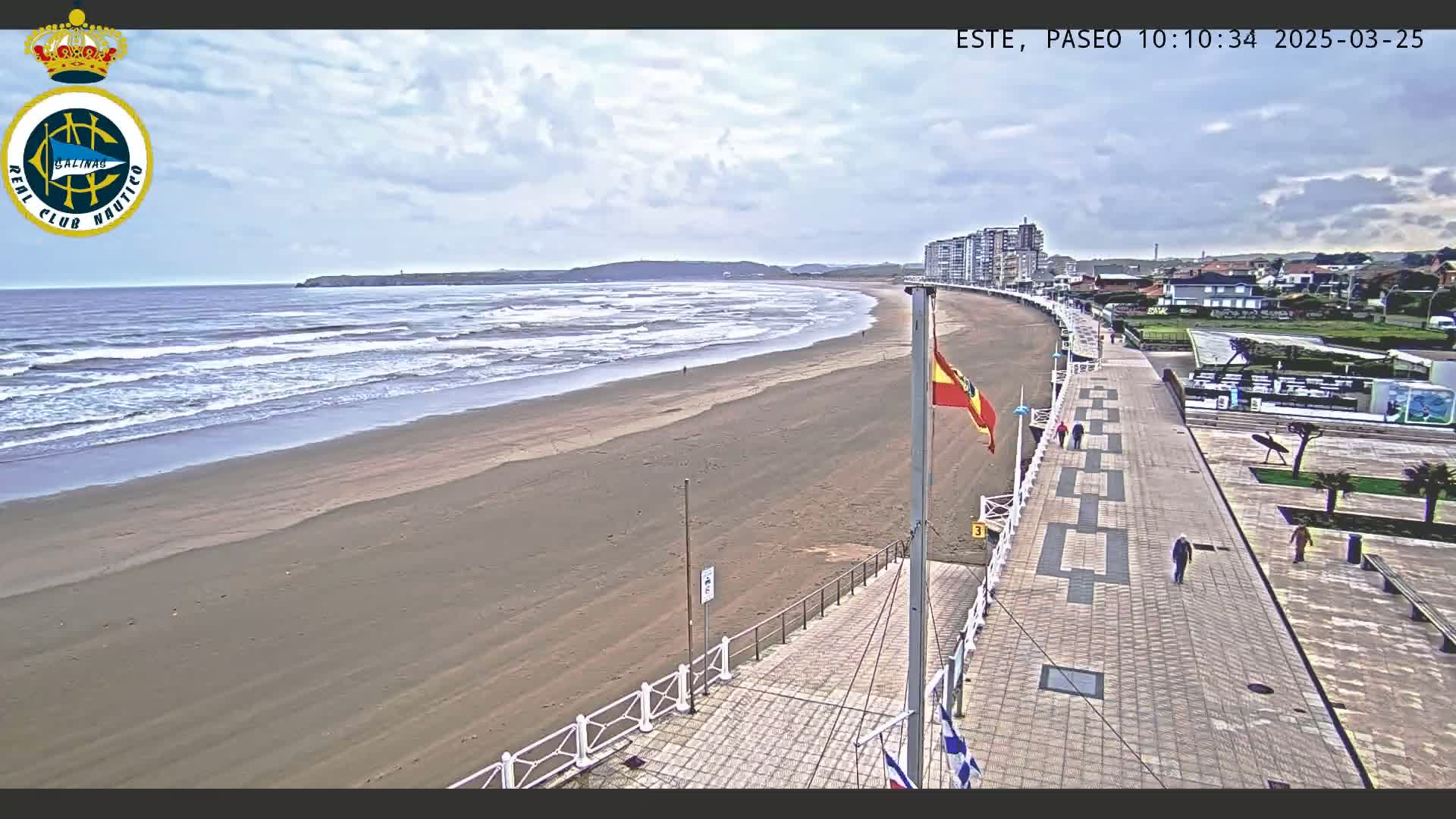A wide sandy beach with waves under a cloudy sky is bordered by a paved walkway and buildings, with a few people strolling along the path.
