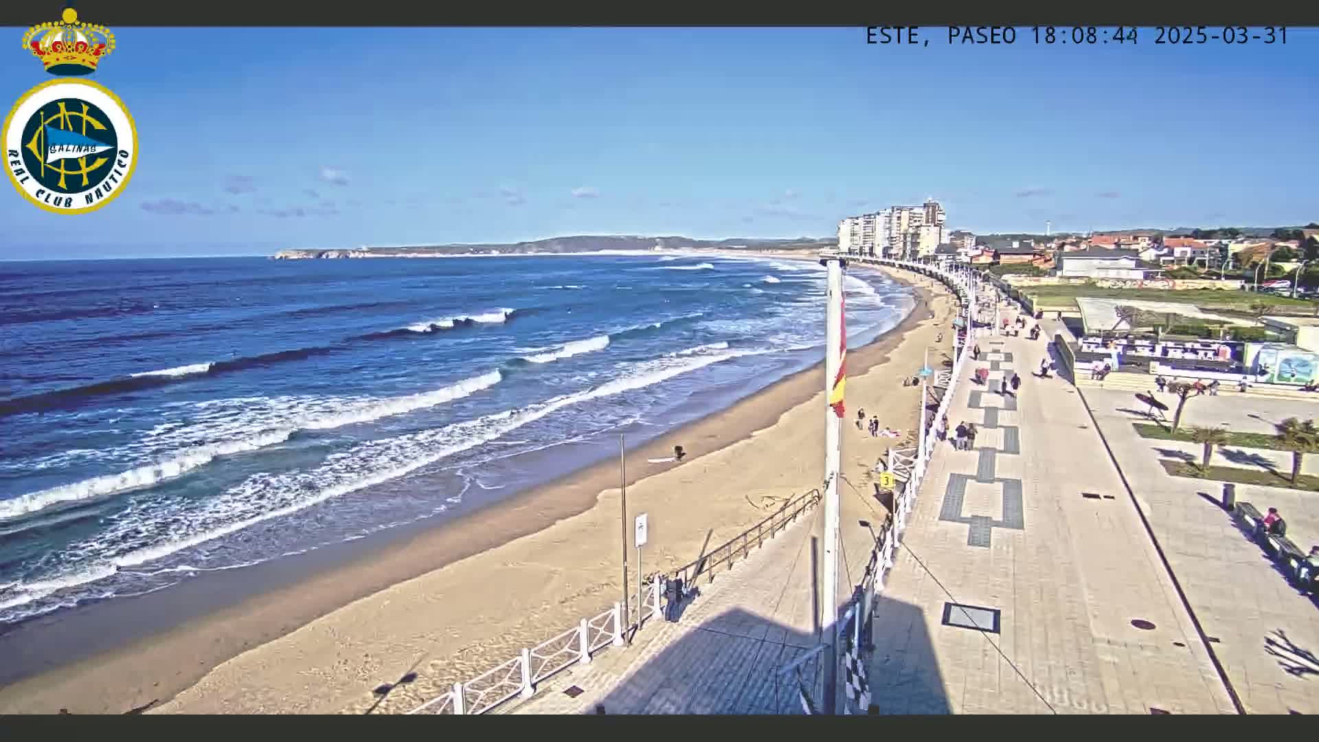 A sandy beach with moderate waves under a clear blue sky is bordered by a paved walkway and buildings.