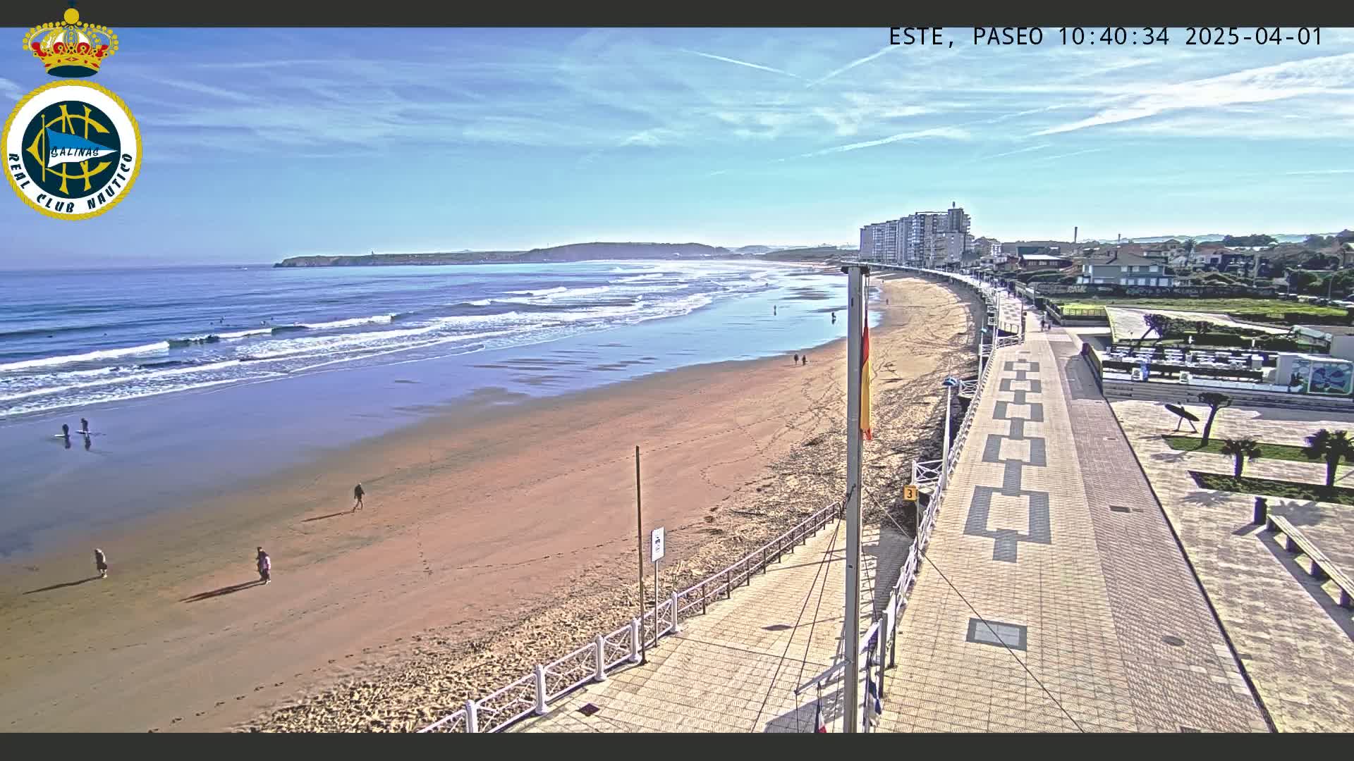 A sandy beach with gentle waves under a clear blue sky is bordered by a paved walkway and buildings.
