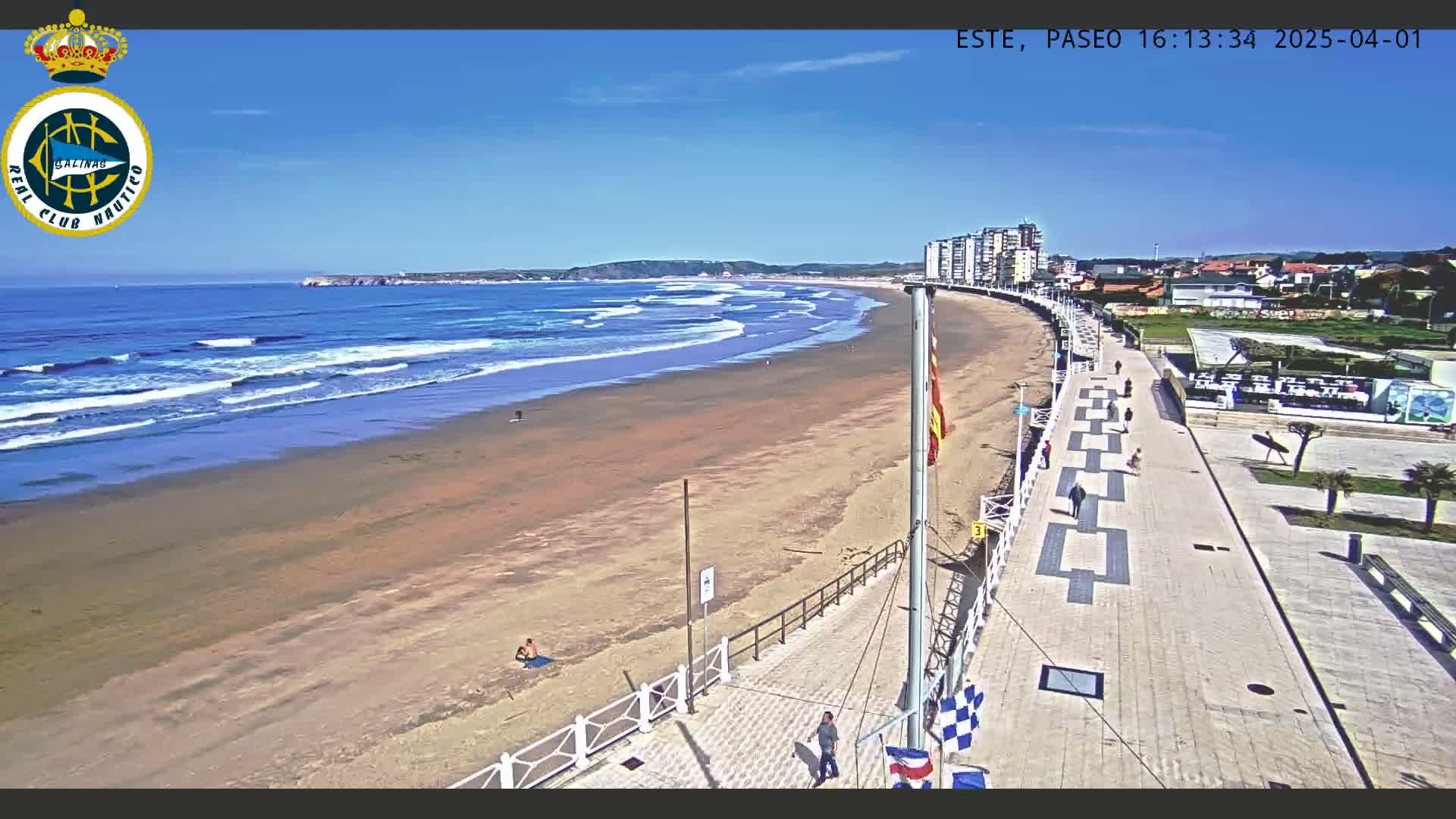 A sandy beach with small waves under a clear blue sky is bordered by a paved walkway with several people strolling, and buildings are visible in the distance.