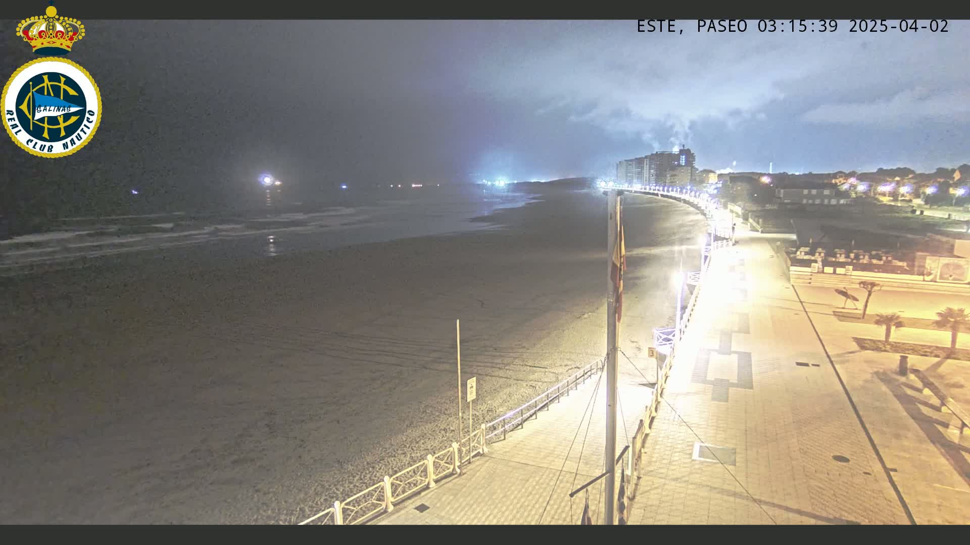 A nighttime view of a mostly empty beach and boardwalk, with calm ocean waves and buildings visible in the distance under a cloudy sky.