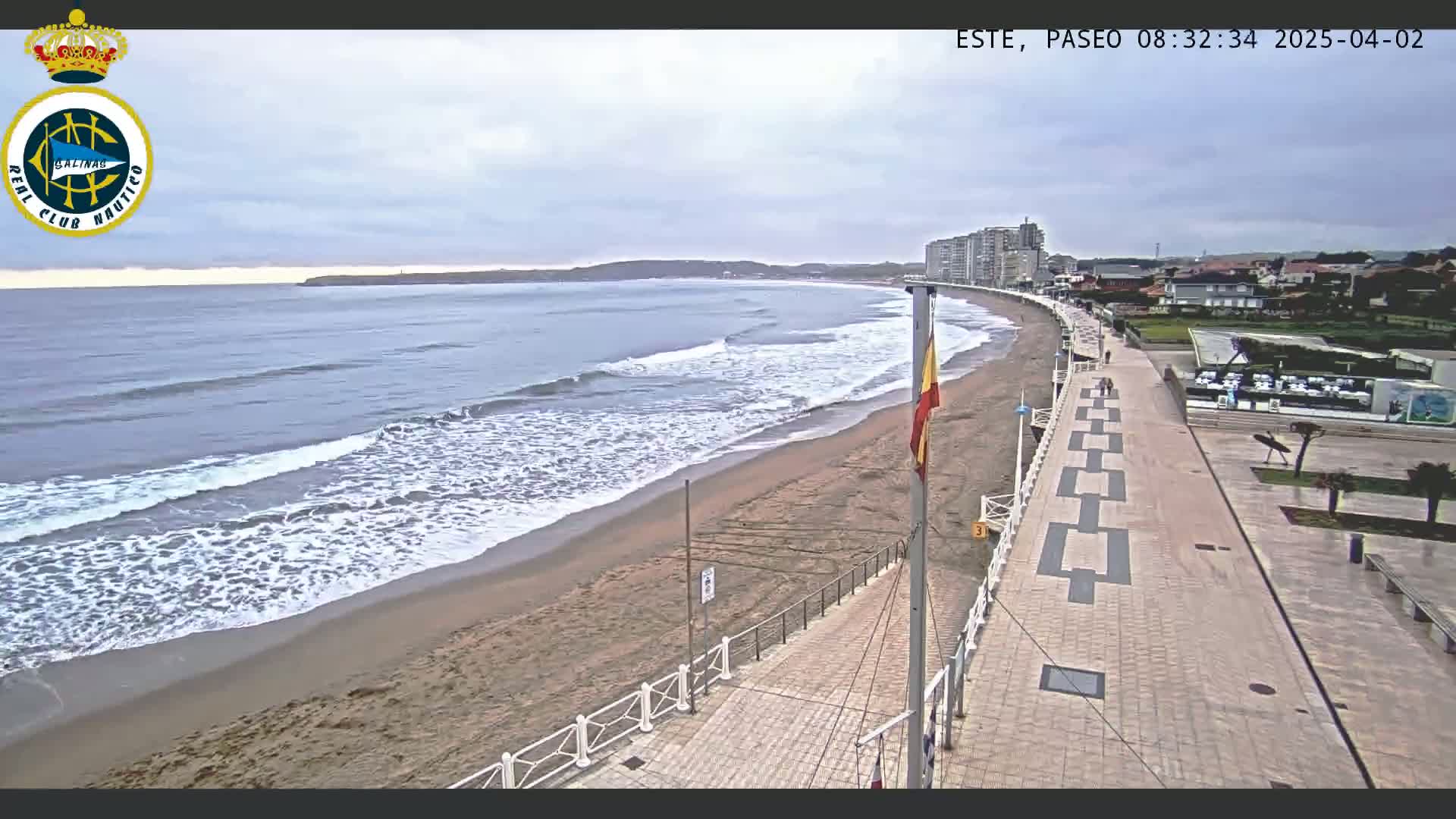 A sandy beach with moderate waves under an overcast sky is bordered by a paved walkway leading to buildings and a parking area.
