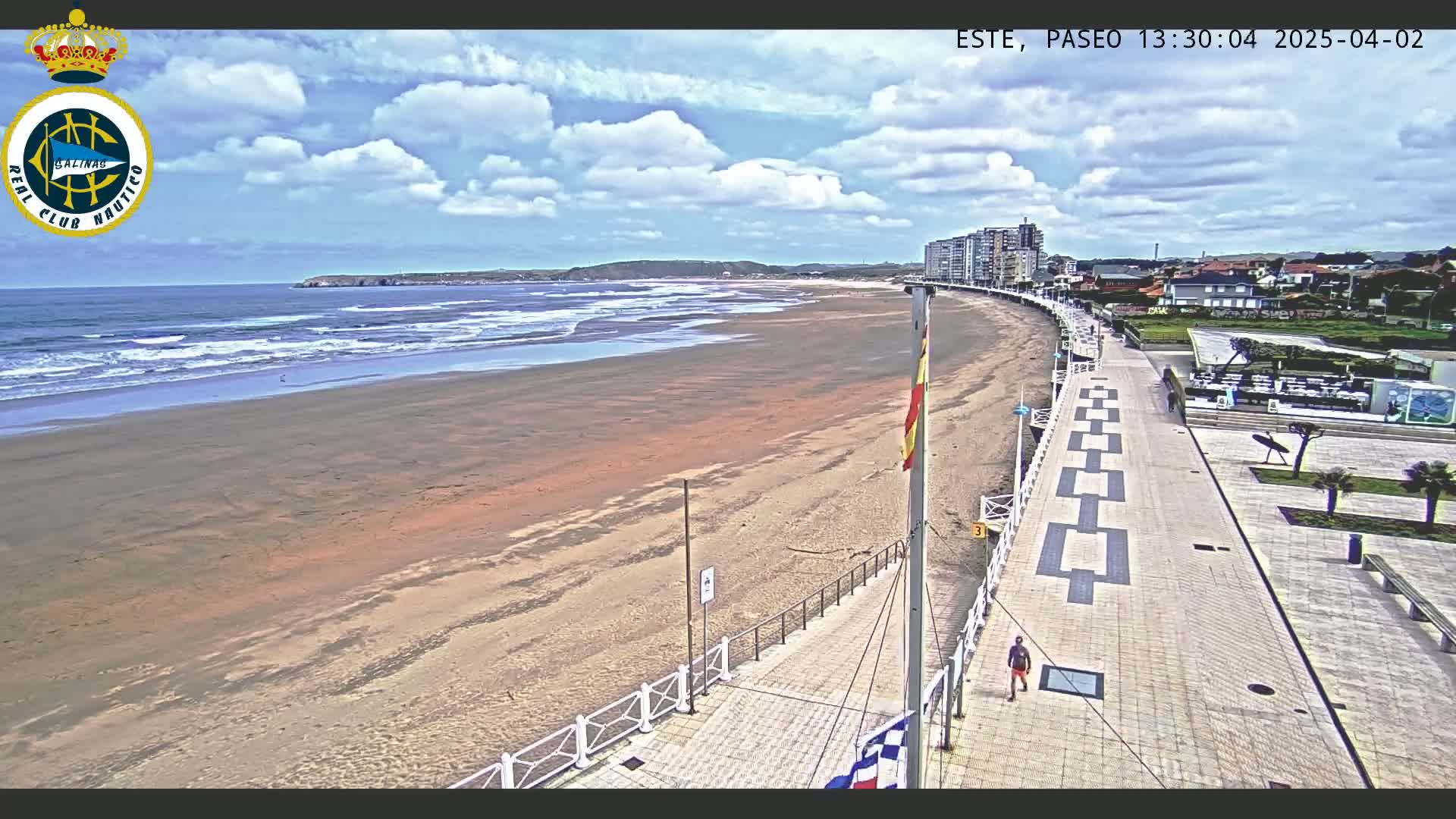 A wide sandy beach with moderate waves under a partly cloudy sky is bordered by a paved walkway and buildings.
