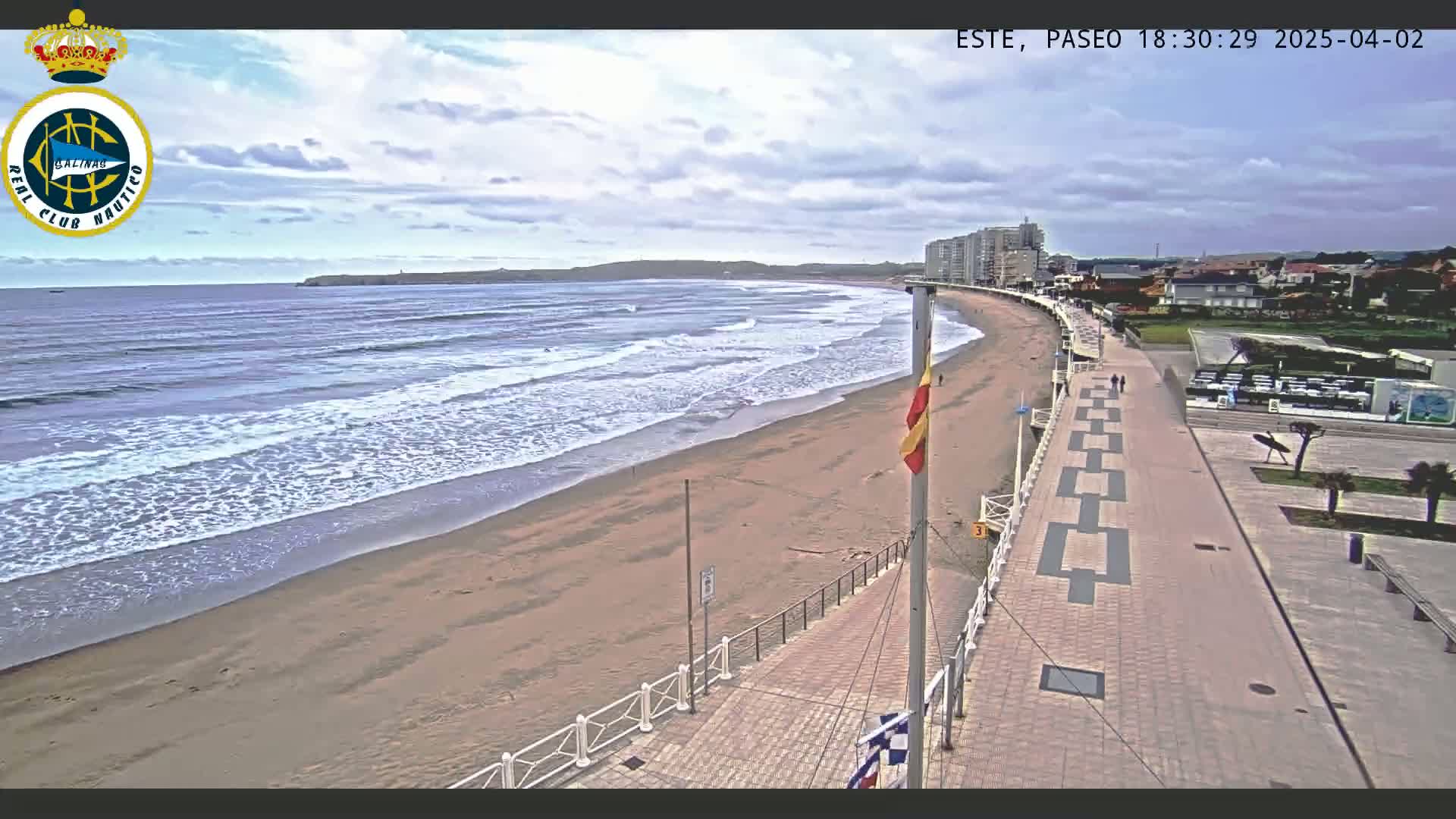 A sandy beach with moderate waves under a partly cloudy sky is bordered by a paved walkway and buildings.