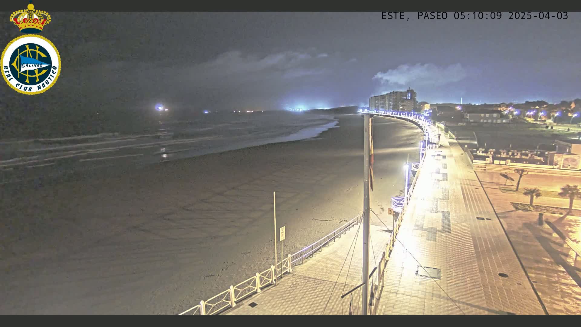 A nighttime view of a mostly empty sandy beach and boardwalk, with calm waves and a few lights visible in the distance under a cloudy sky.
