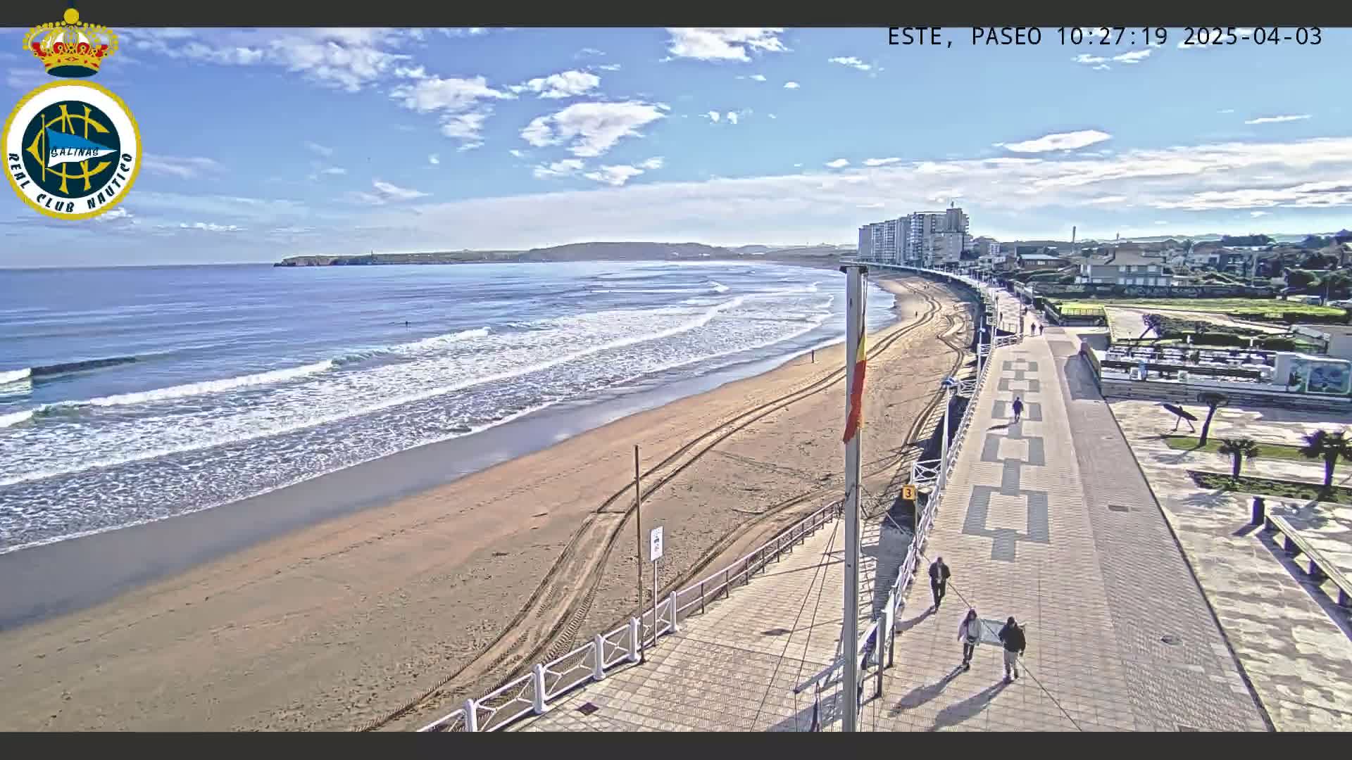 A sandy beach with gentle waves under a mostly sunny sky is adjacent to a paved boardwalk with several people walking, and buildings are visible in the distance.