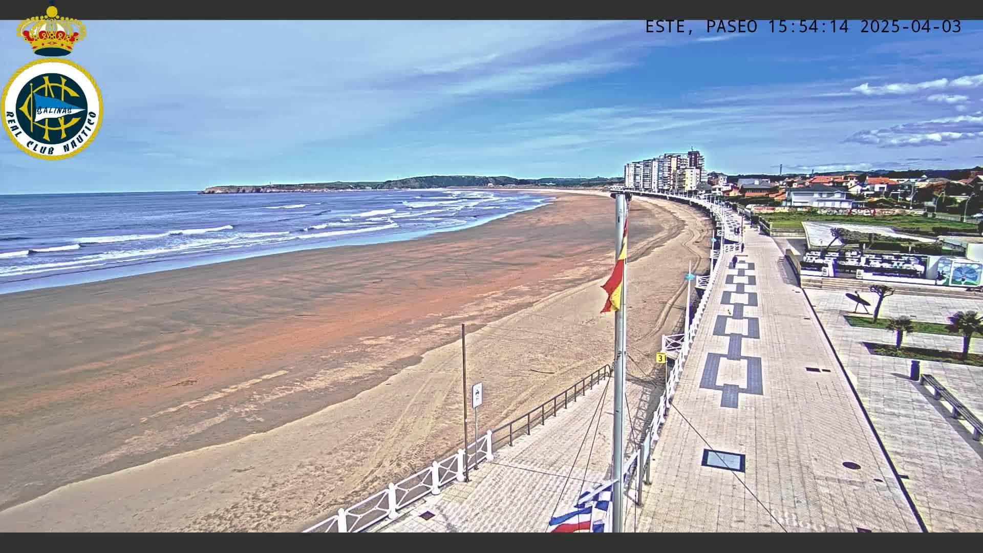 A long sandy beach with small waves under a mostly clear sky is adjacent to a paved boardwalk and seaside buildings.