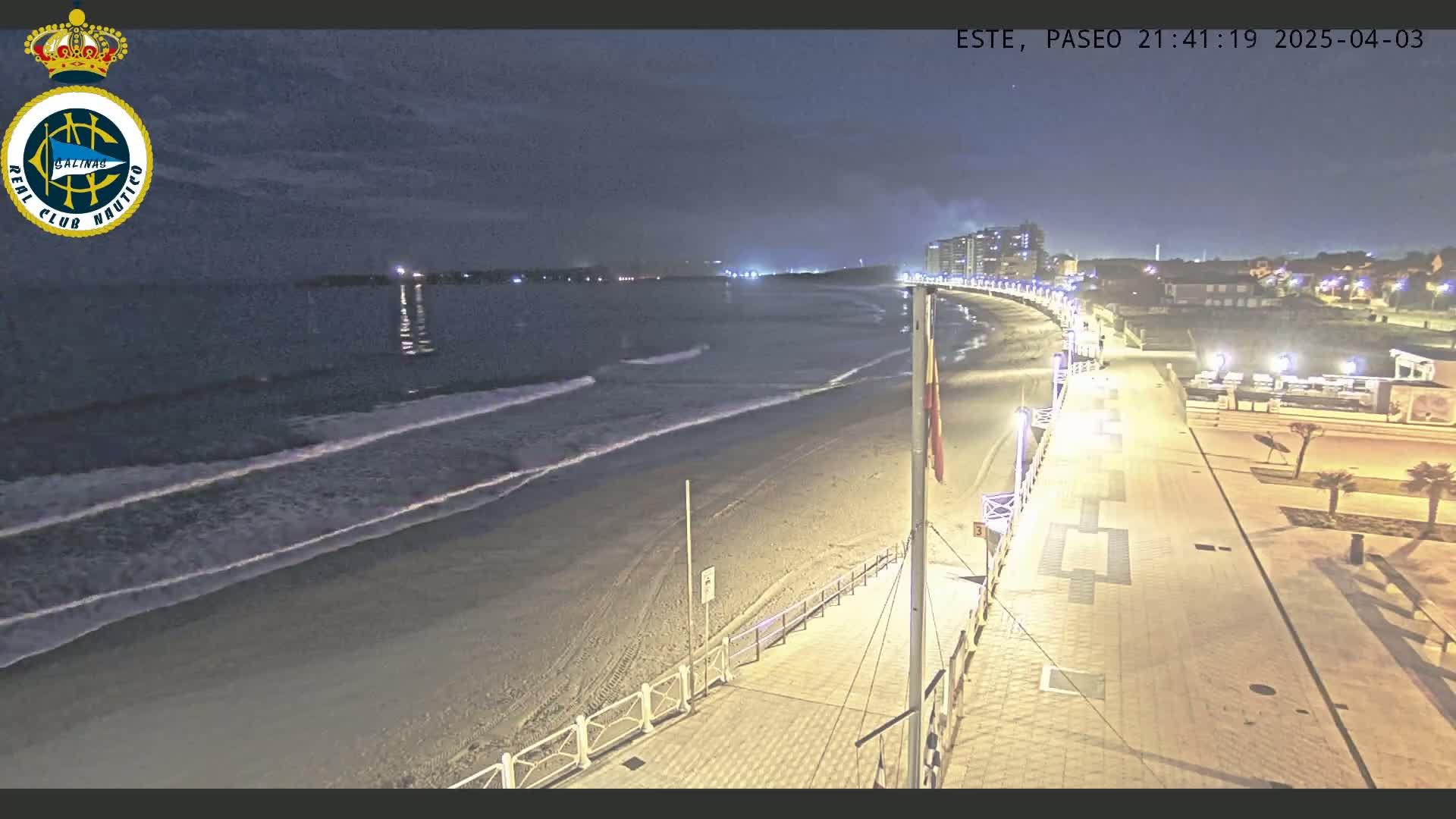 A nighttime view of a sandy beach with gentle waves lapping the shore, a boardwalk lined with lights, and buildings in the distance under a mostly clear sky.