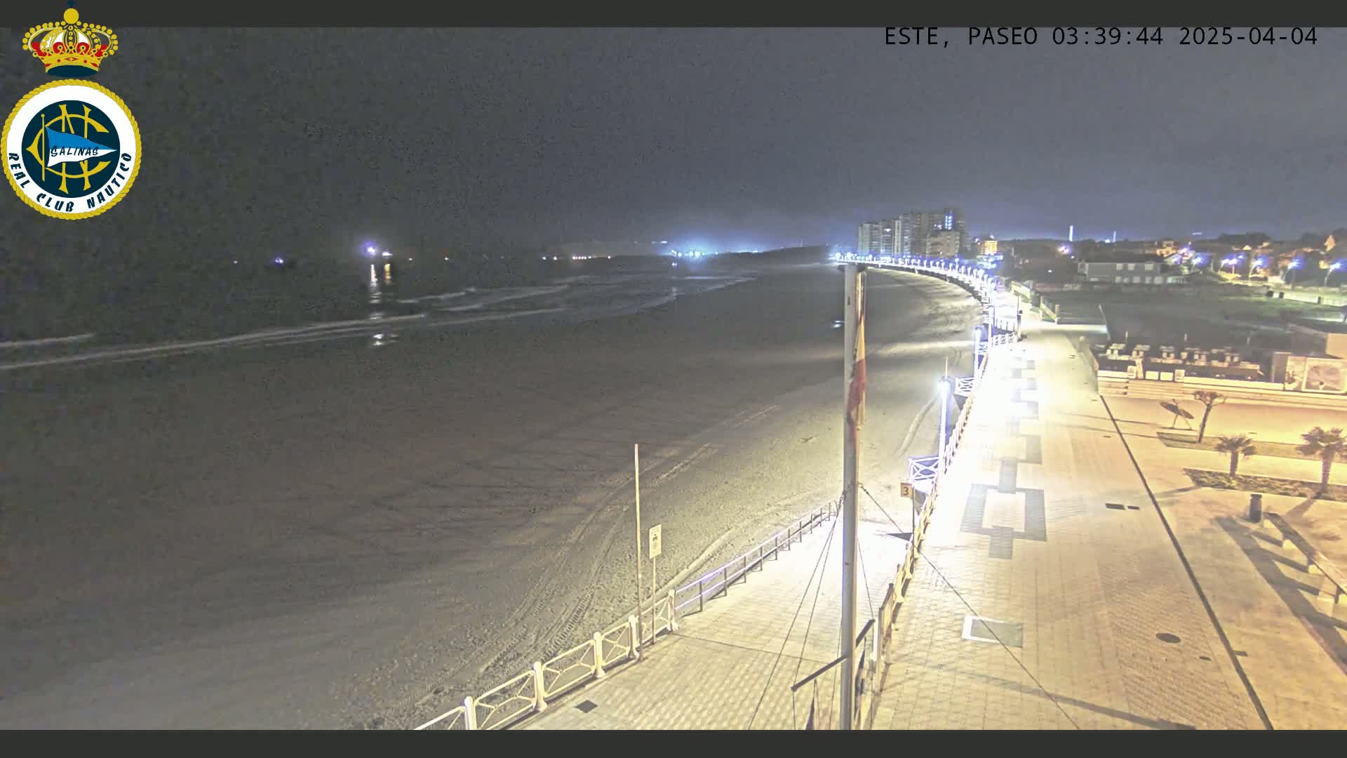 A dark beach at night is shown, with a paved walkway and buildings in the background, lit by streetlights.