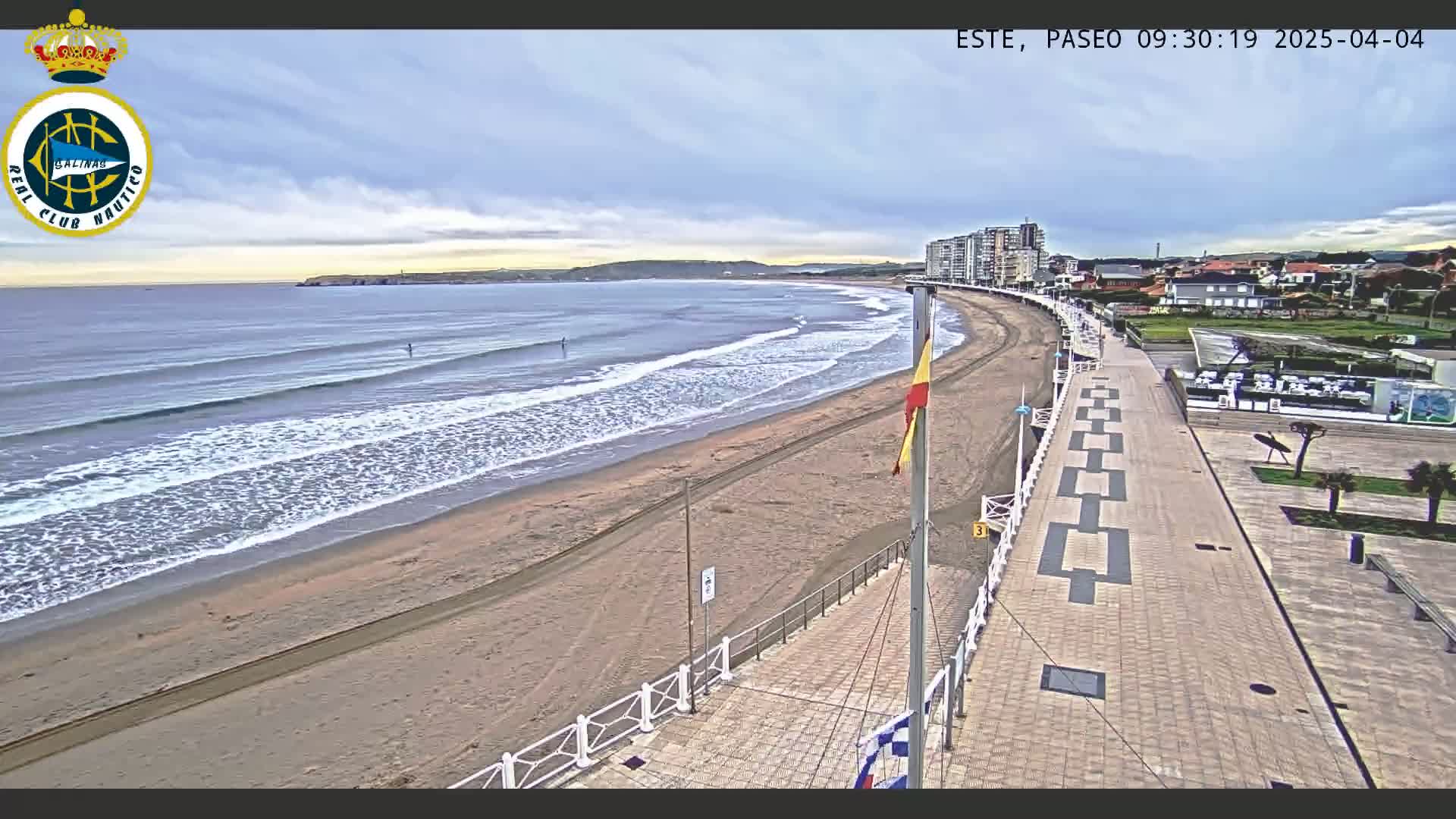 A sandy beach with gentle waves under a partly cloudy sky is bordered by a paved promenade and buildings.