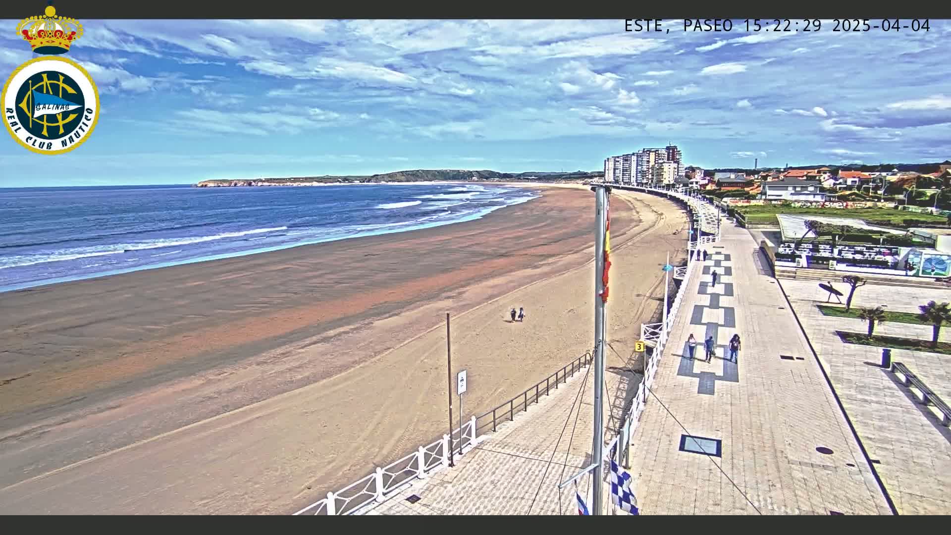 A wide sandy beach with gentle waves under a partly cloudy blue sky is bordered by a paved walkway with a few pedestrians, leading to buildings and a town.