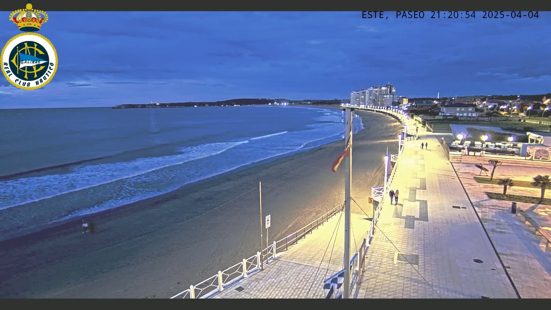 A beach at twilight, with gentle waves lapping the shore, is bordered by a brightly lit promenade with a few pedestrians.