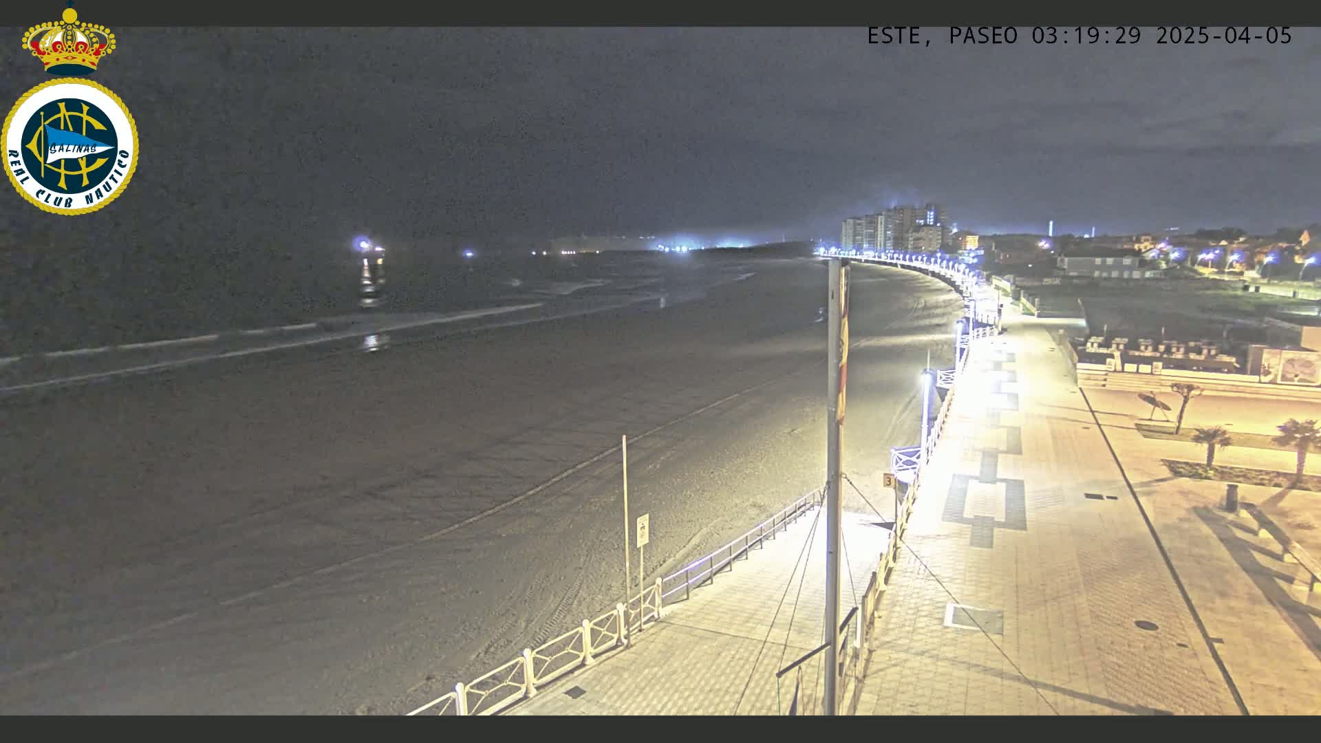 A nighttime, high-angle view shows a mostly empty, dark beach with a paved walkway and some buildings in the distance.