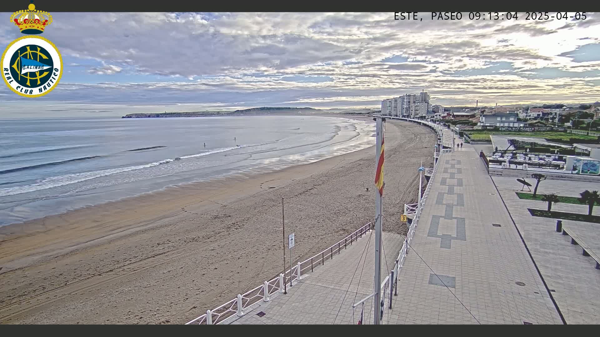 A mostly cloudy day reveals a sandy beach with gentle waves, a paved boardwalk lined with buildings, and a few people strolling along the shore.