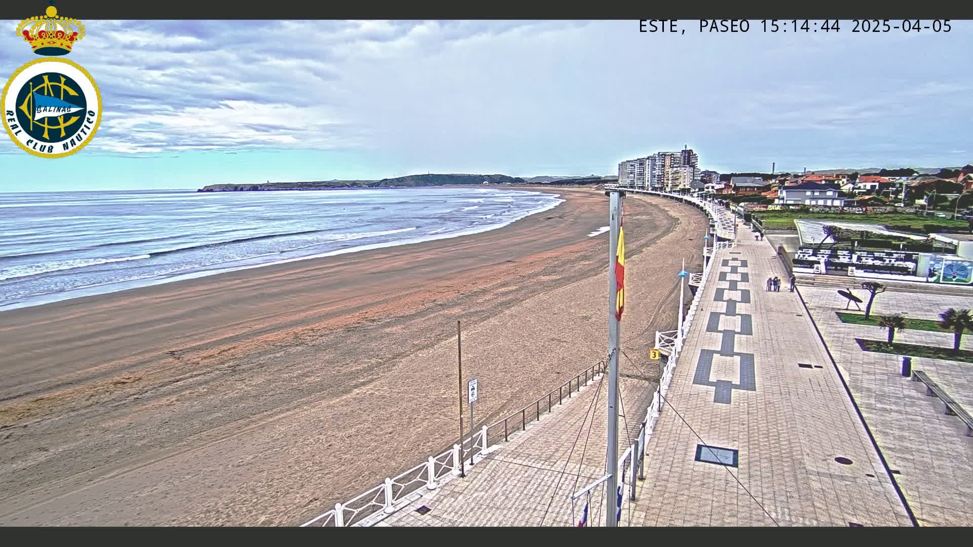 A wide sandy beach with gentle waves stretches along a paved promenade lined with buildings under a mostly cloudy sky.